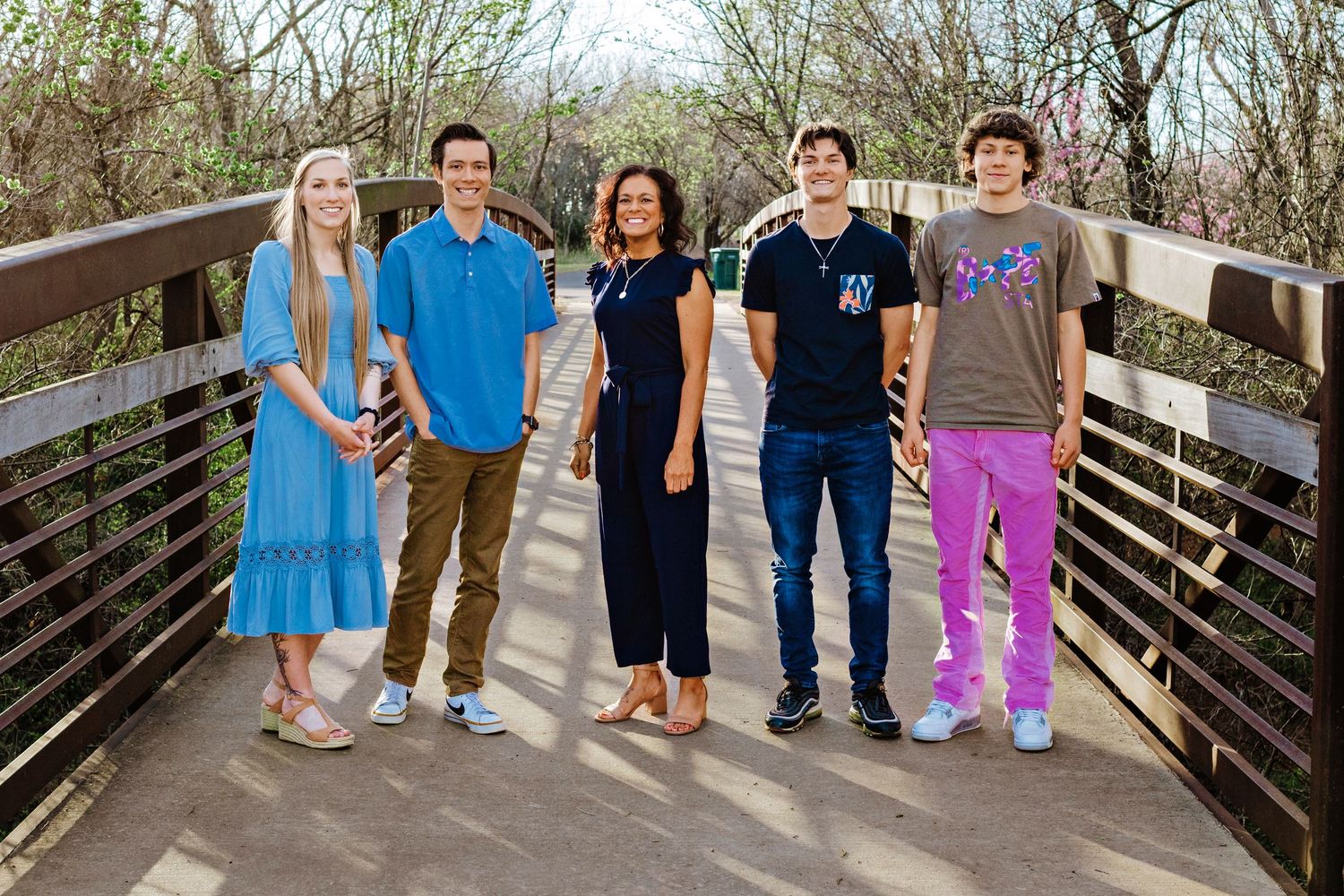 A group of five people standing together on a wooden bridge walkway surrounded by trees in casual attire.