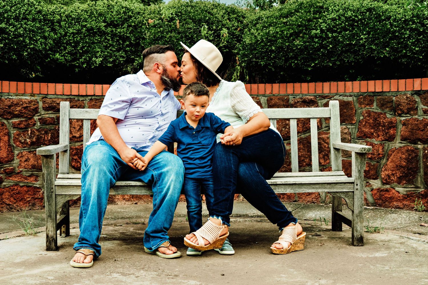 Family of three sitting on wooden bench, mother kissing fathers cheek with young son between them outdoors.