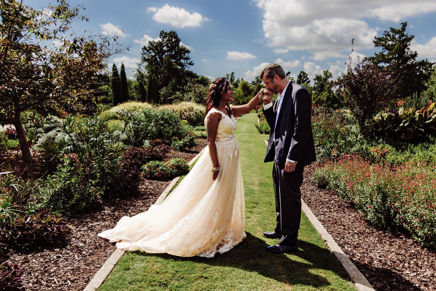 Bride and groom share a tender moment in a lush garden, with the bride in a flowing white gown touching the groom's face.