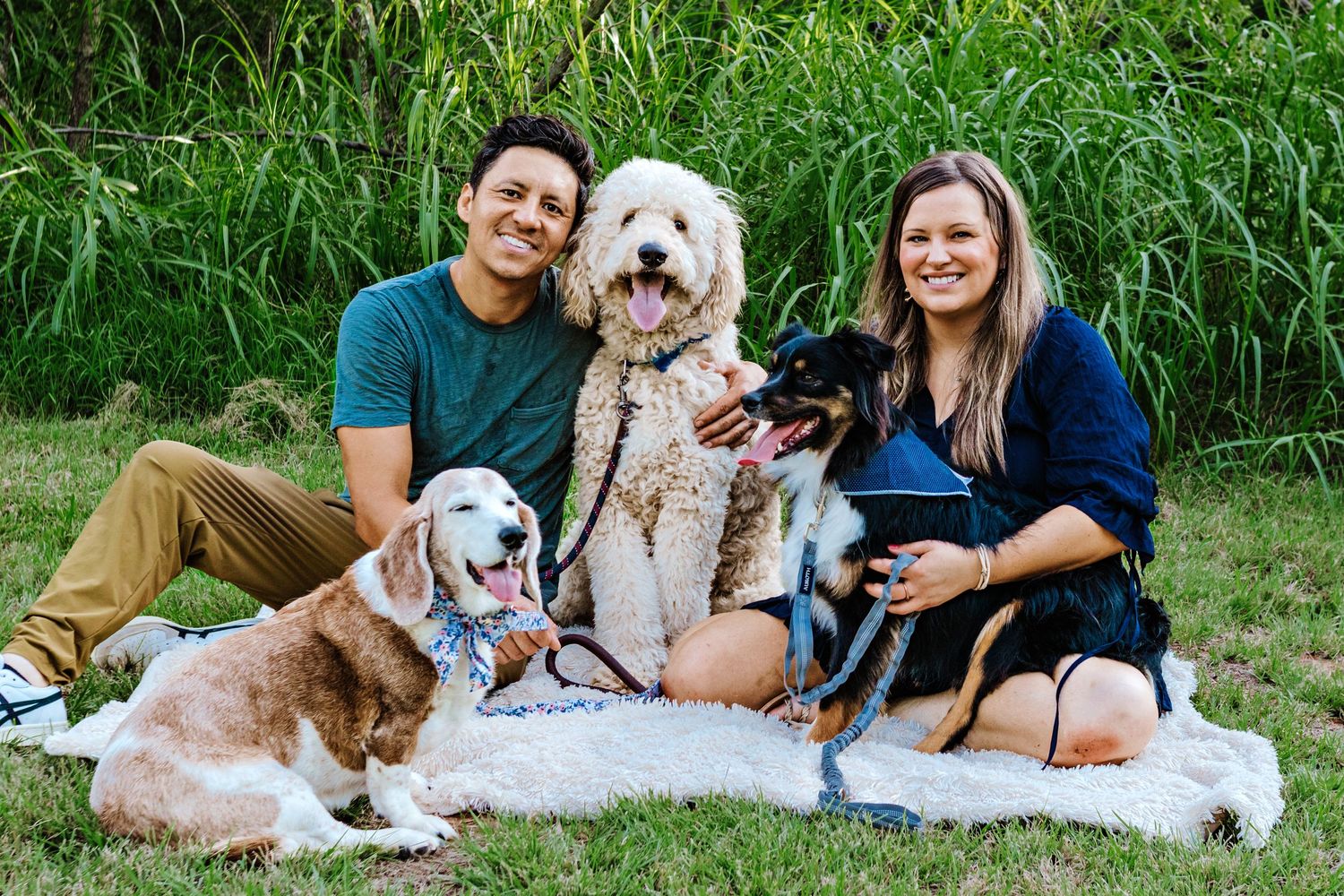A couple sits on a blanket outdoors with their three dogs including a doodle and two smaller breeds.