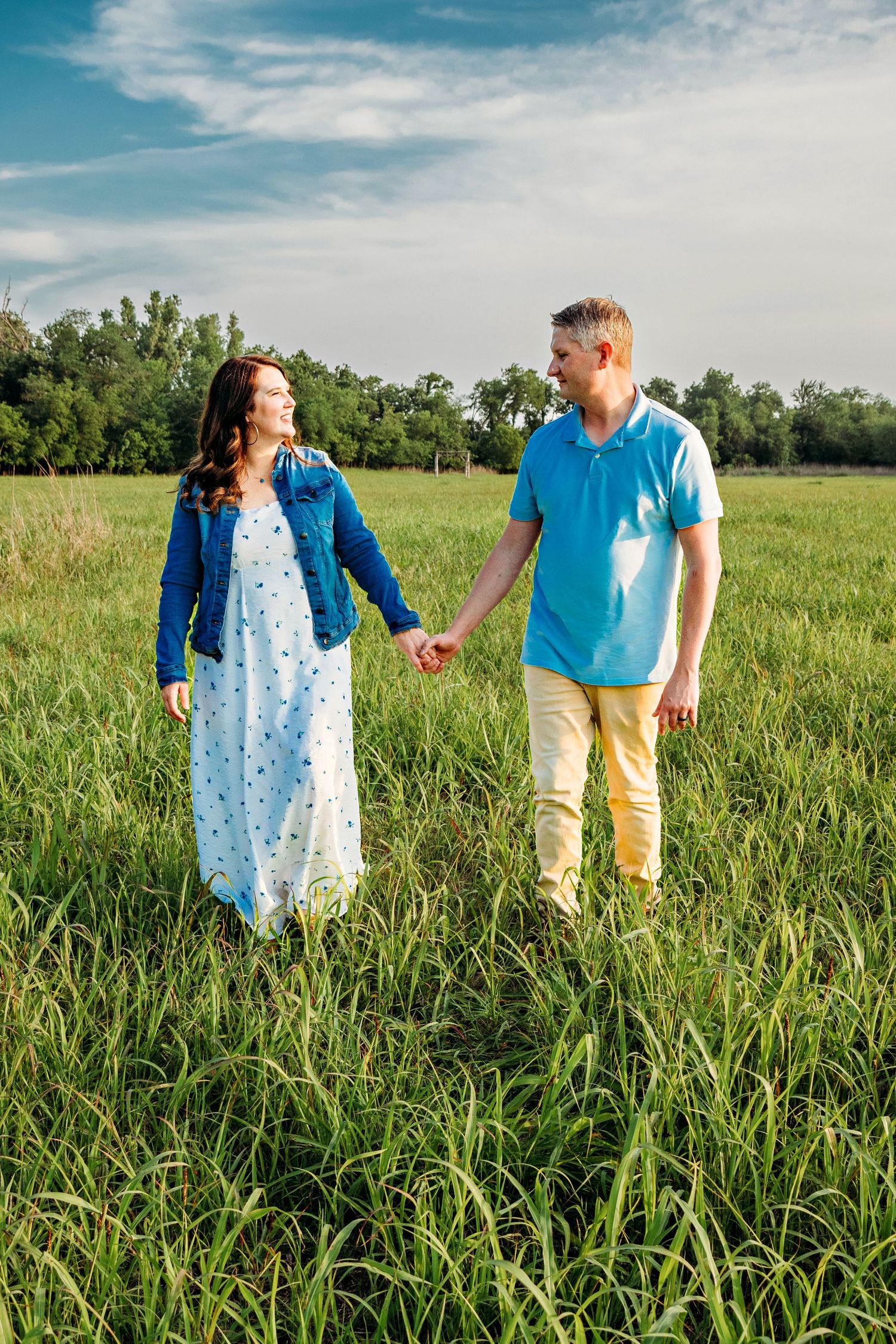 A couple in blue and yellow outfits holds hands while walking through a lush green field on a sunny summer day.