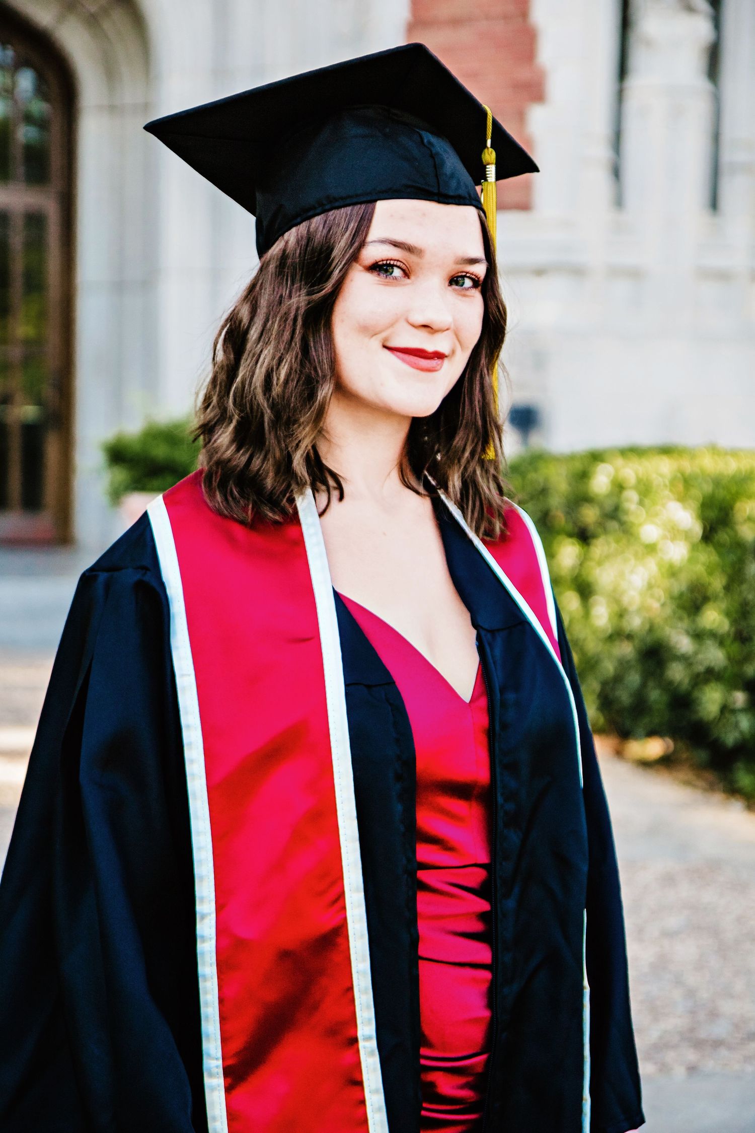 Graduate in black cap and gown with red stole poses for portrait series outdoors at commencement ceremony.