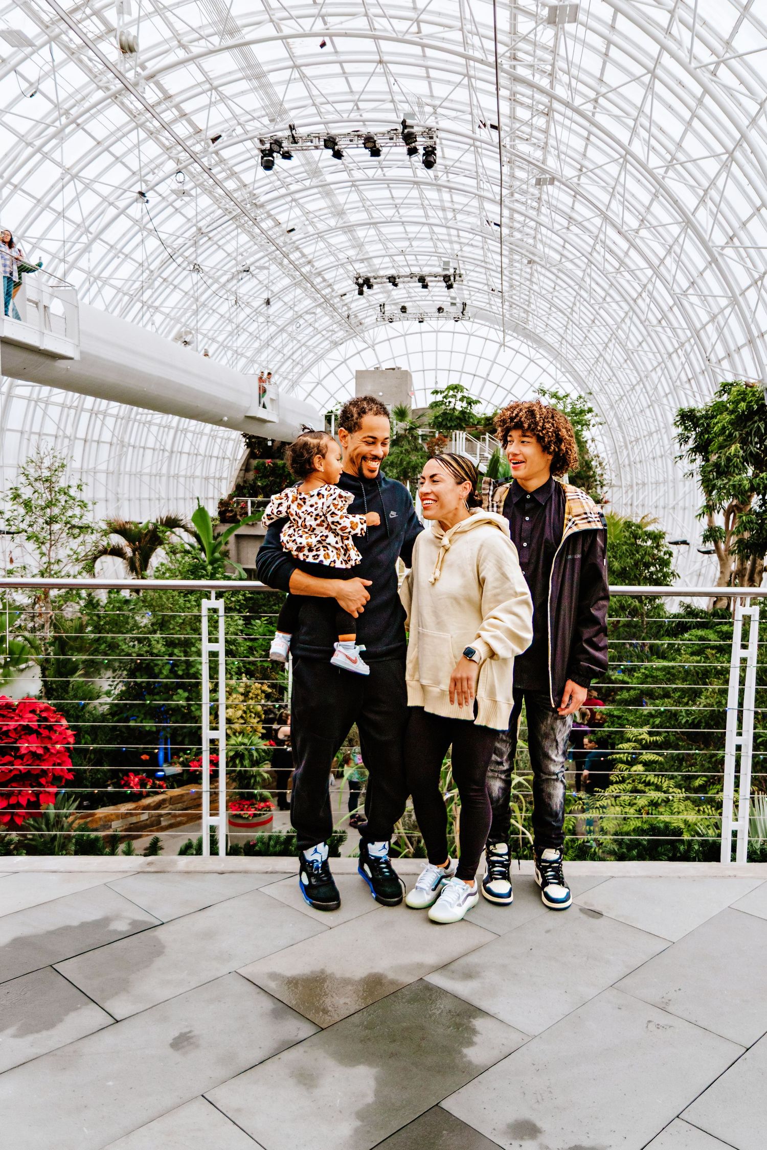 A joyful family of four poses inside a glass-domed botanical garden, with lush greenery in the background.