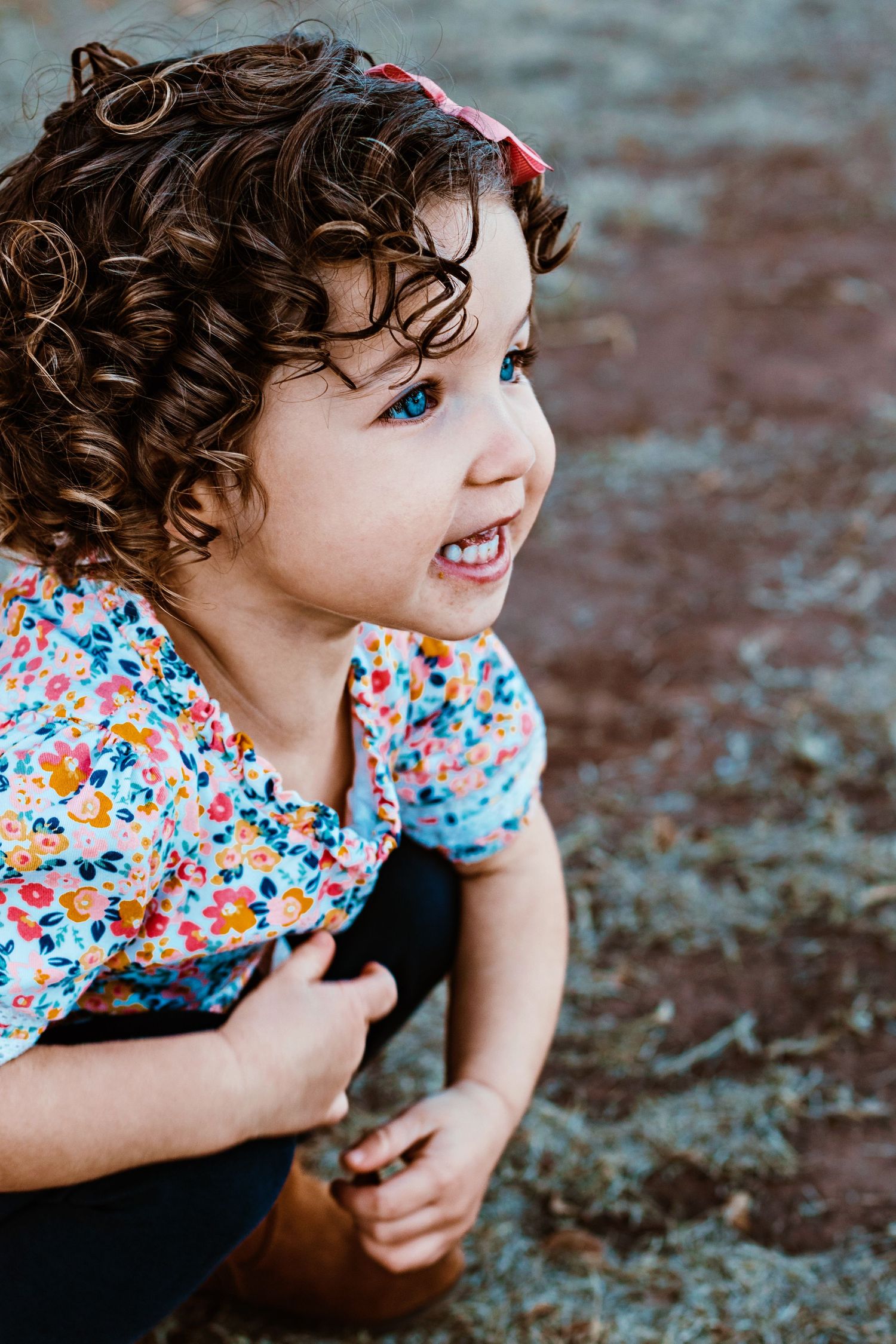 Child in floral print dress smiles and plays outdoors with curly hair and joyful expression.