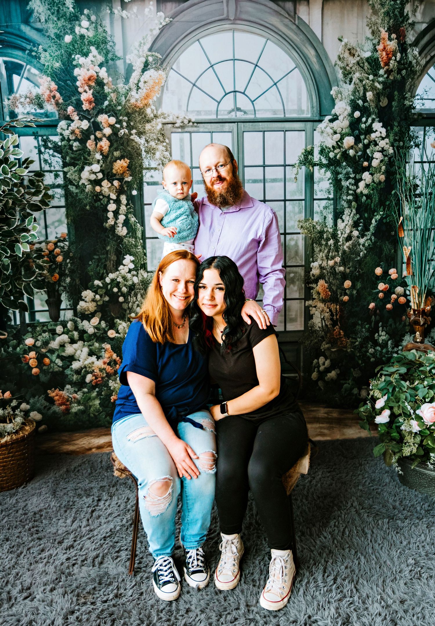 A family group poses together in front of an elegant arched window surrounded by flowers.