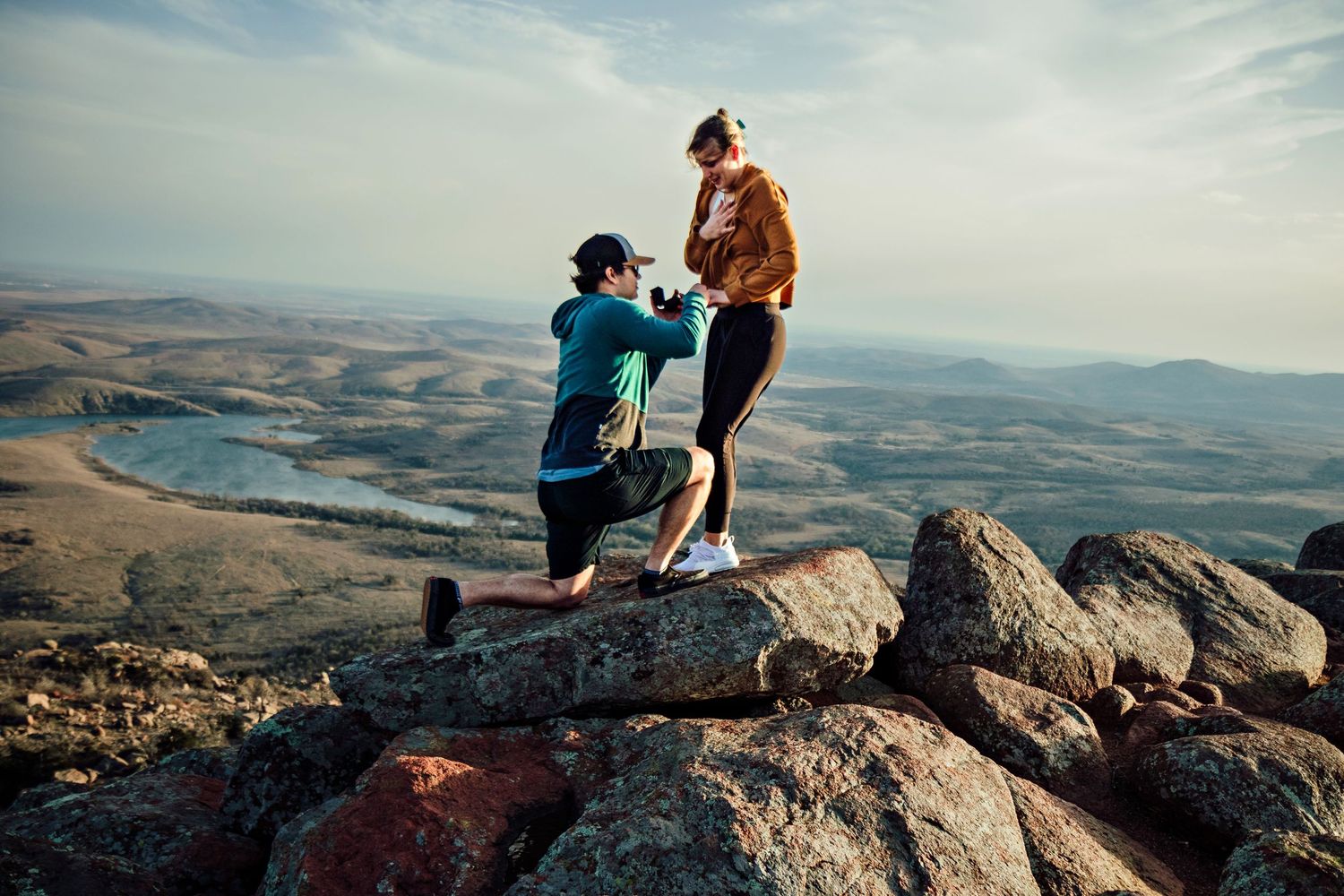 A couple shares a romantic moment during sunset at a mountain summit overlooking a vast landscape.