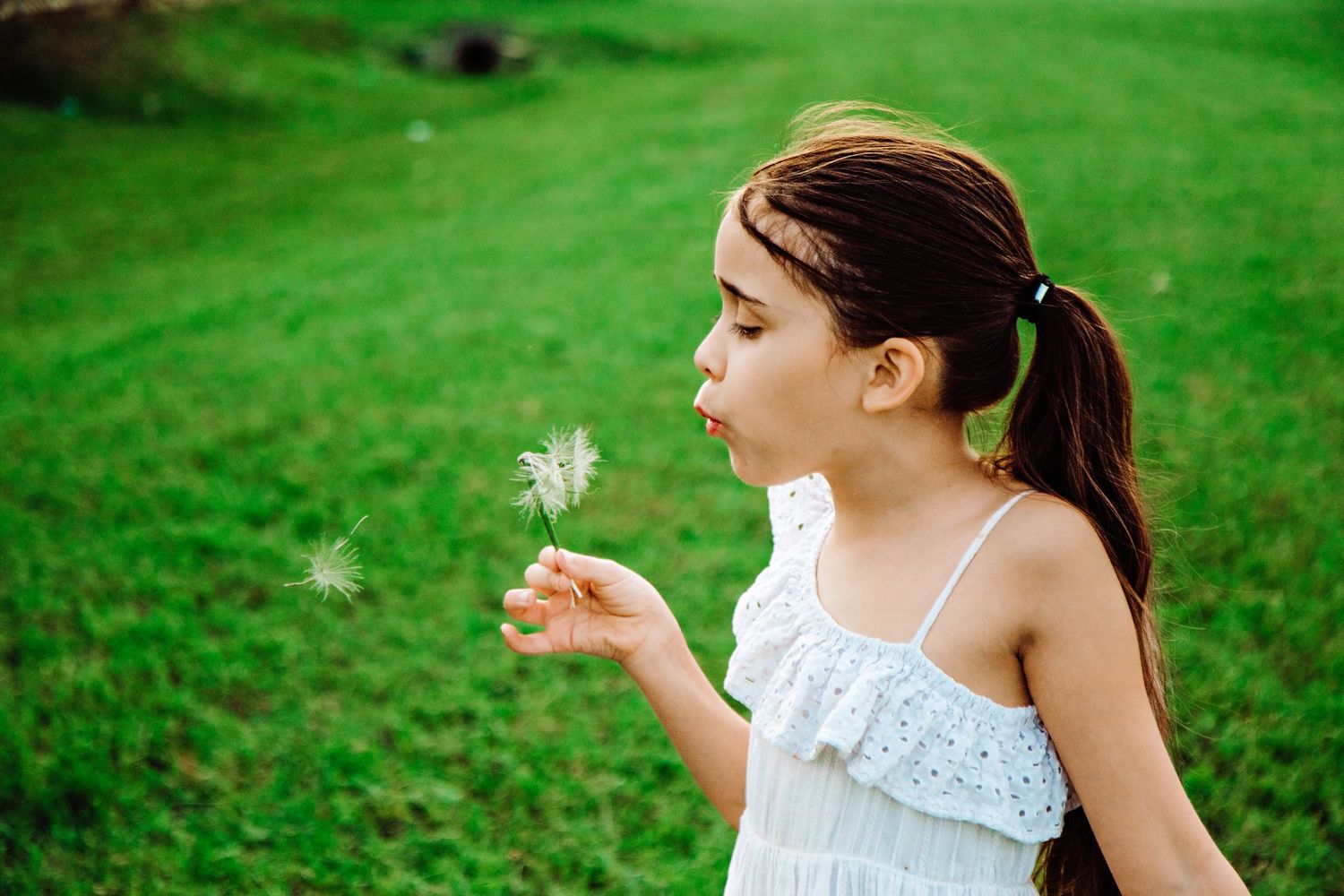 A young girl in a white dress blows dandelion seeds in a lush green field.