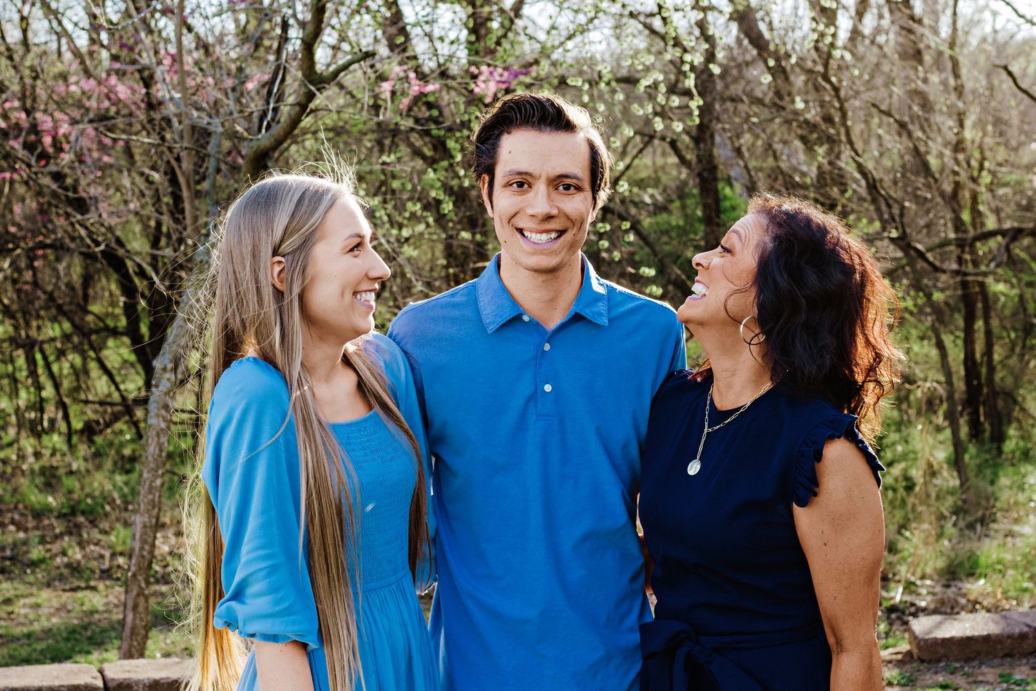 Three people in blue and navy outfits share a joyful moment together outdoors with flowering trees in the background.