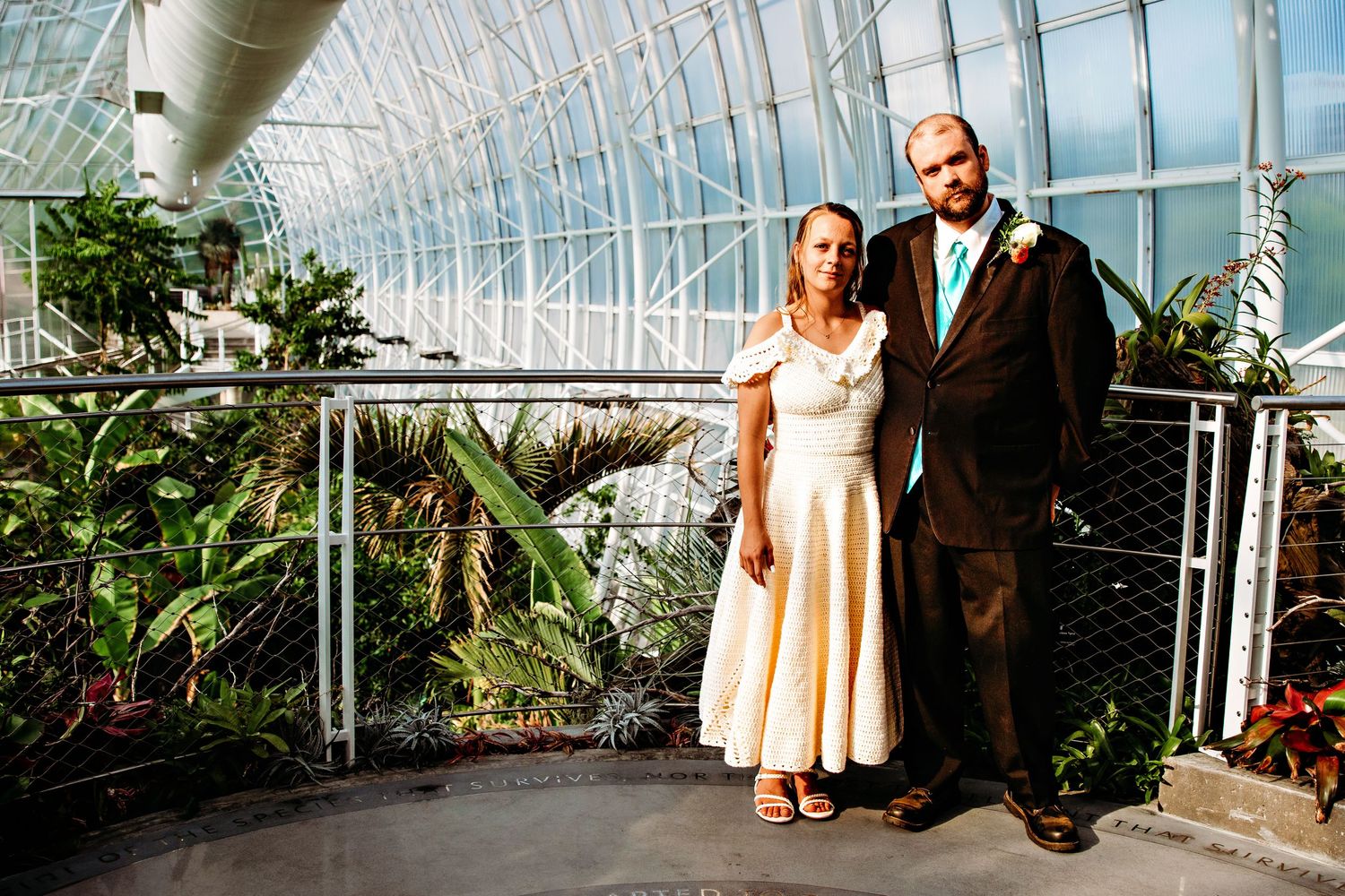 Two people stand together inside a bright greenhouse conservatory with glass ceilings and tropical plants.