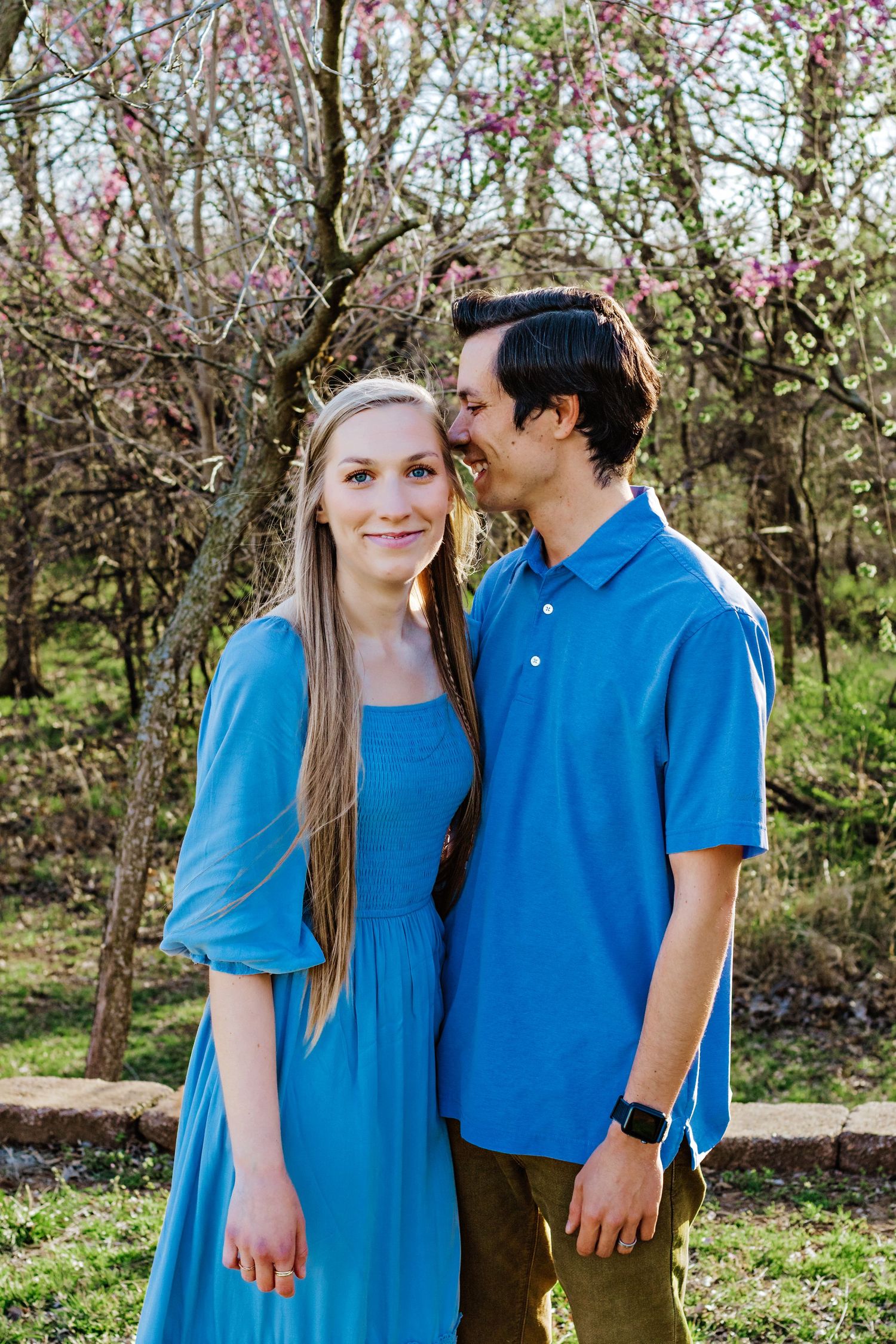 A couple in coordinating blue outfits share a tender moment under blooming spring trees in a garden setting.