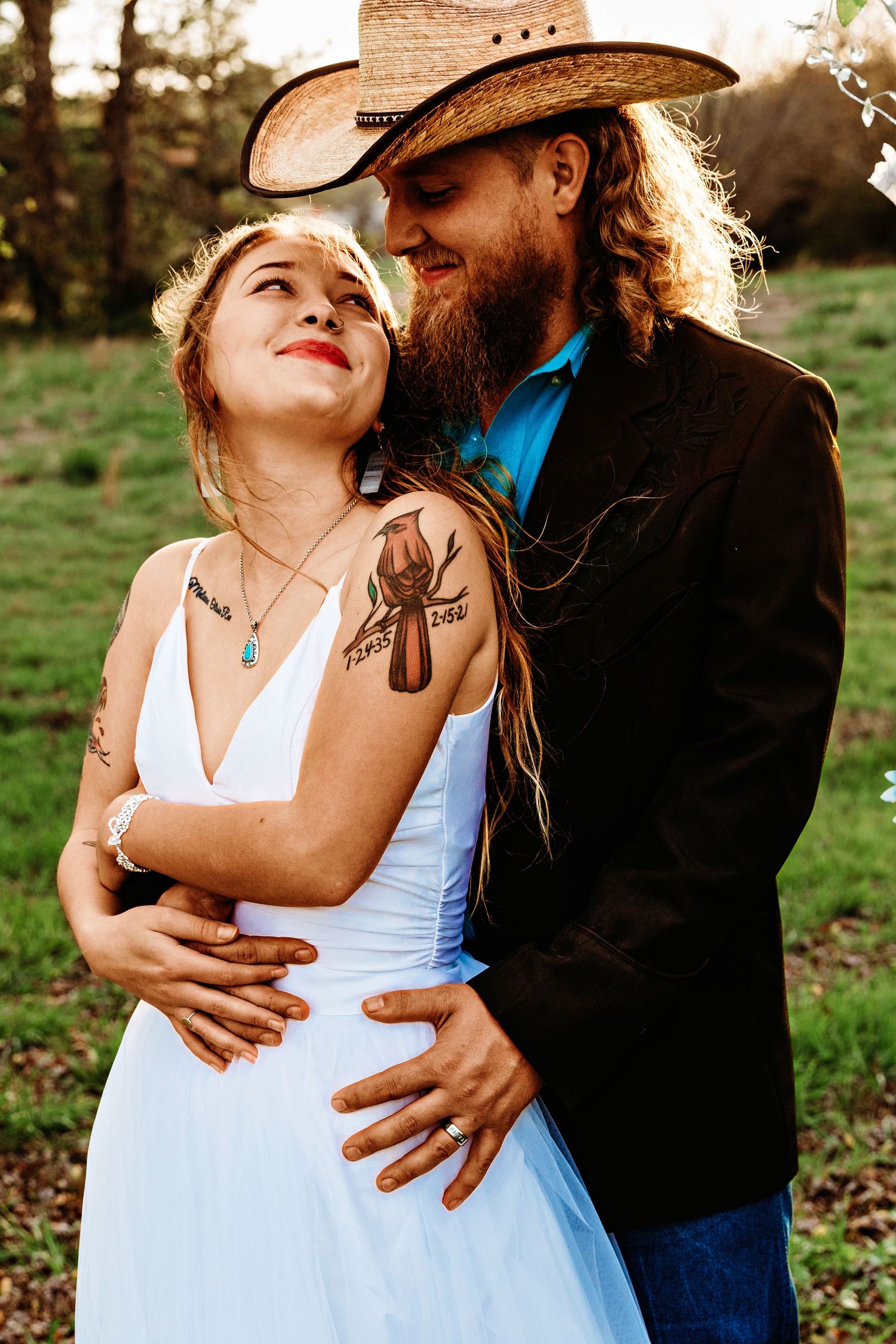 A couple shares an intimate moment outdoors, with one wearing a white dress and the other in denim and a cowboy hat.