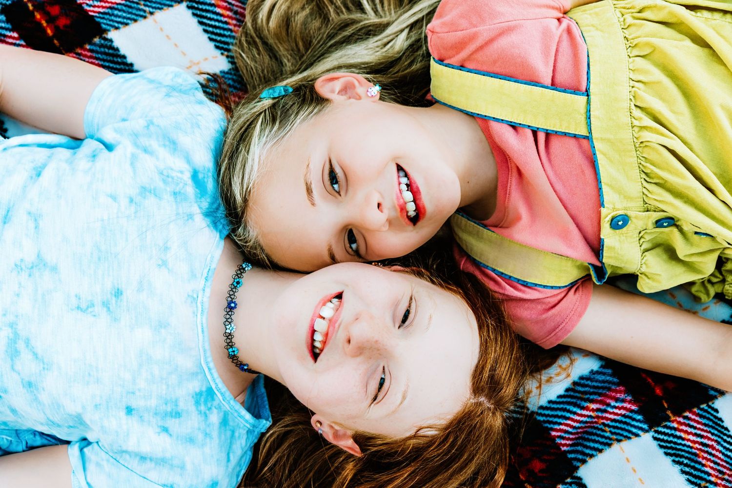 Two smiling girls lying on a colorful plaid blanket, looking up at the camera on a sunny day.