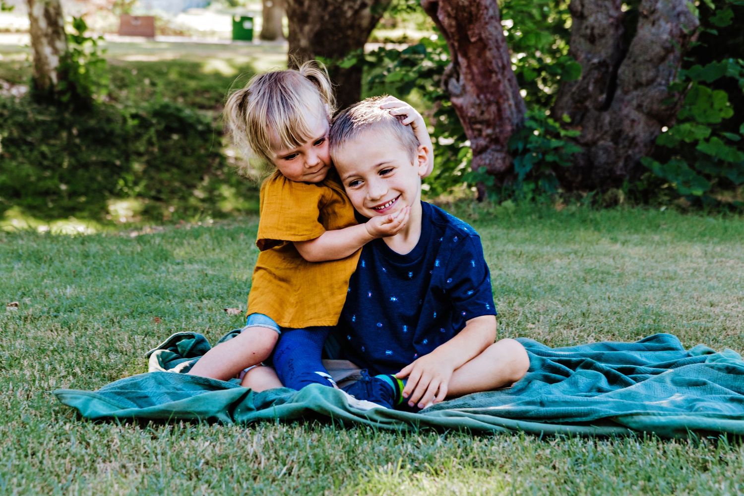 Two young boys sitting on a teal blanket in a grassy park, smiling and playing together outdoors.