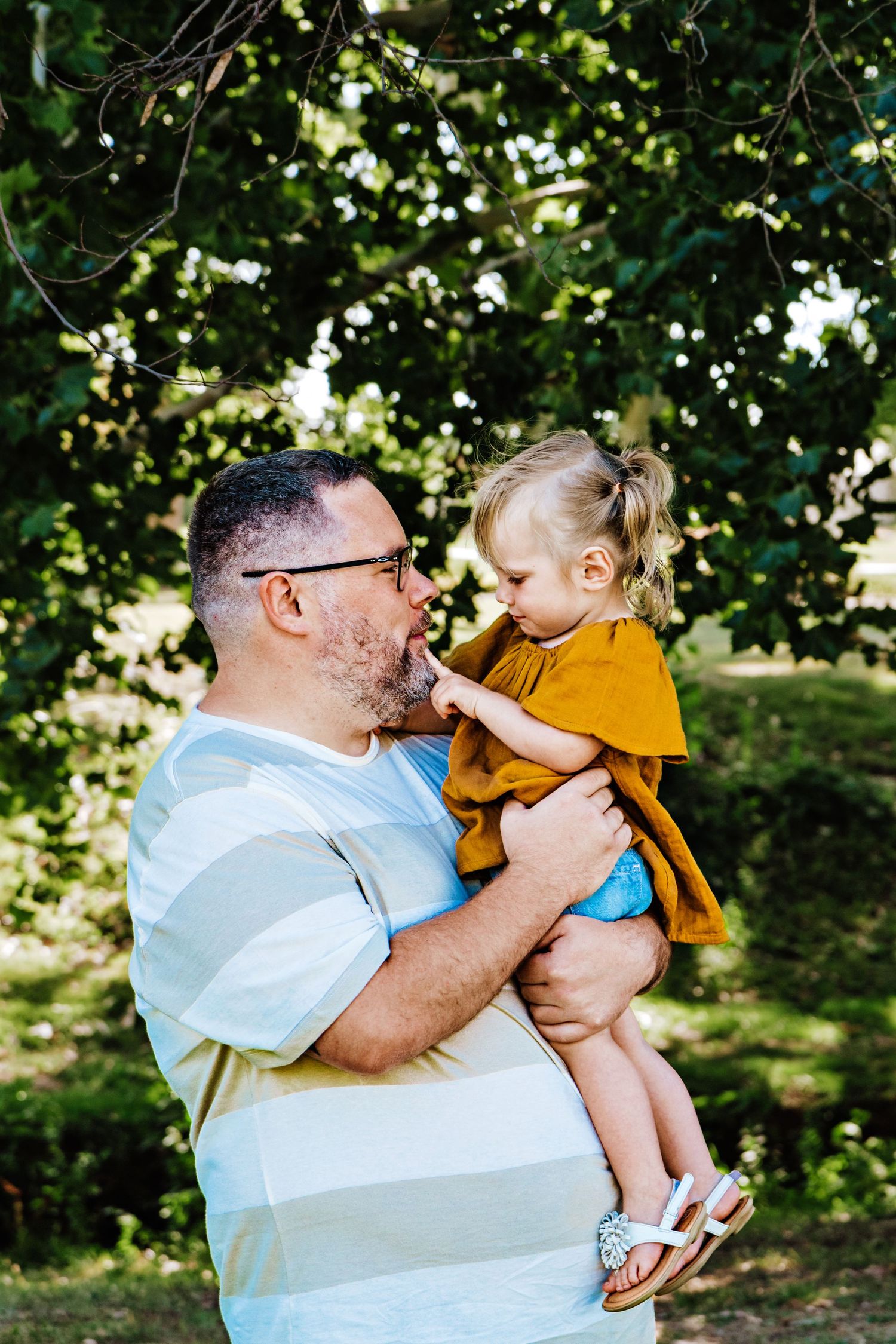 A father holding his young toddler daughter outdoors, sharing a tender moment surrounded by lush green trees.