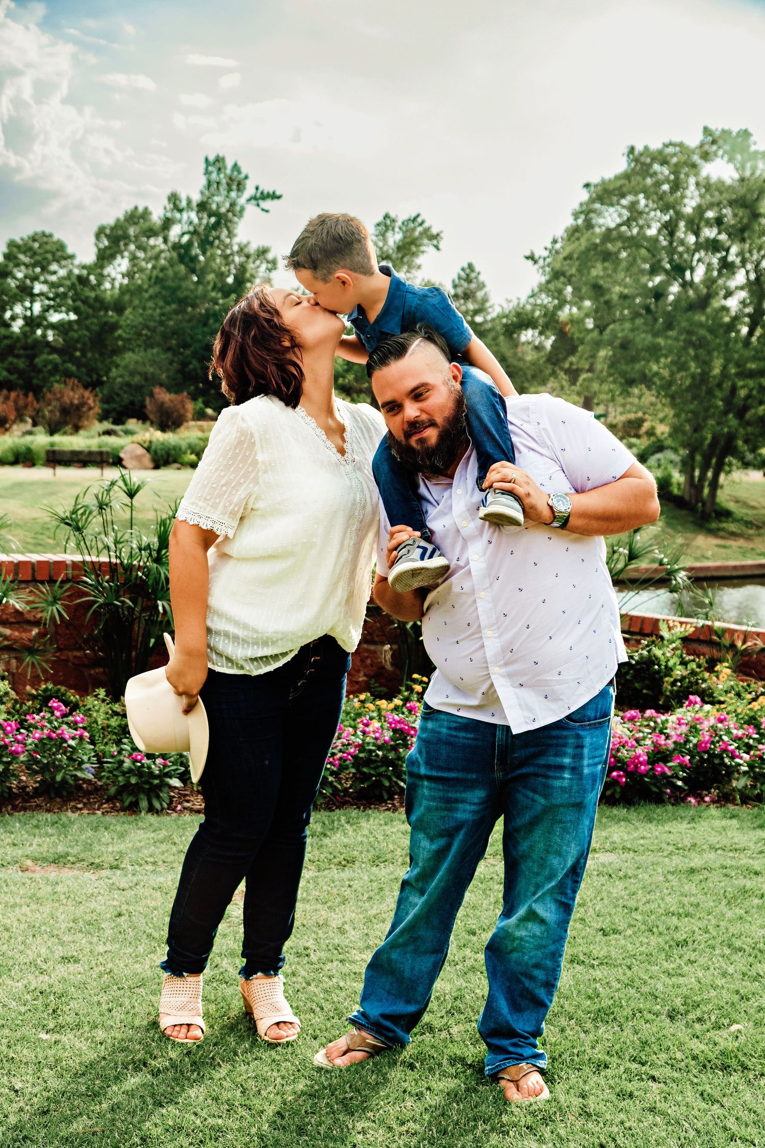 Happy family portrait with parents kissing toddler son outdoors in lush garden with colorful flowers.