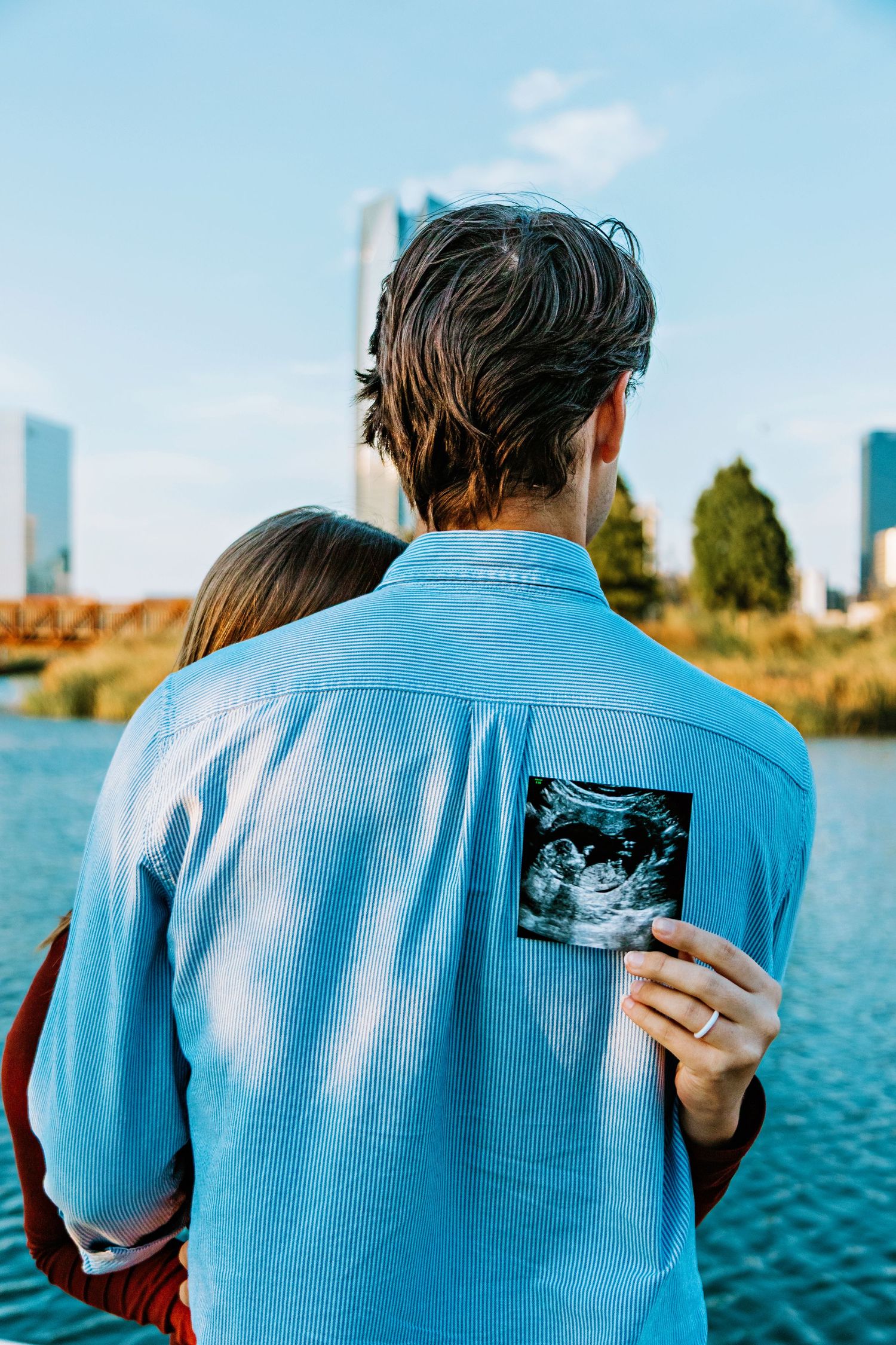 Person in blue shirt holds ultrasound photo against their back while standing near waterfront with city skyline in background.