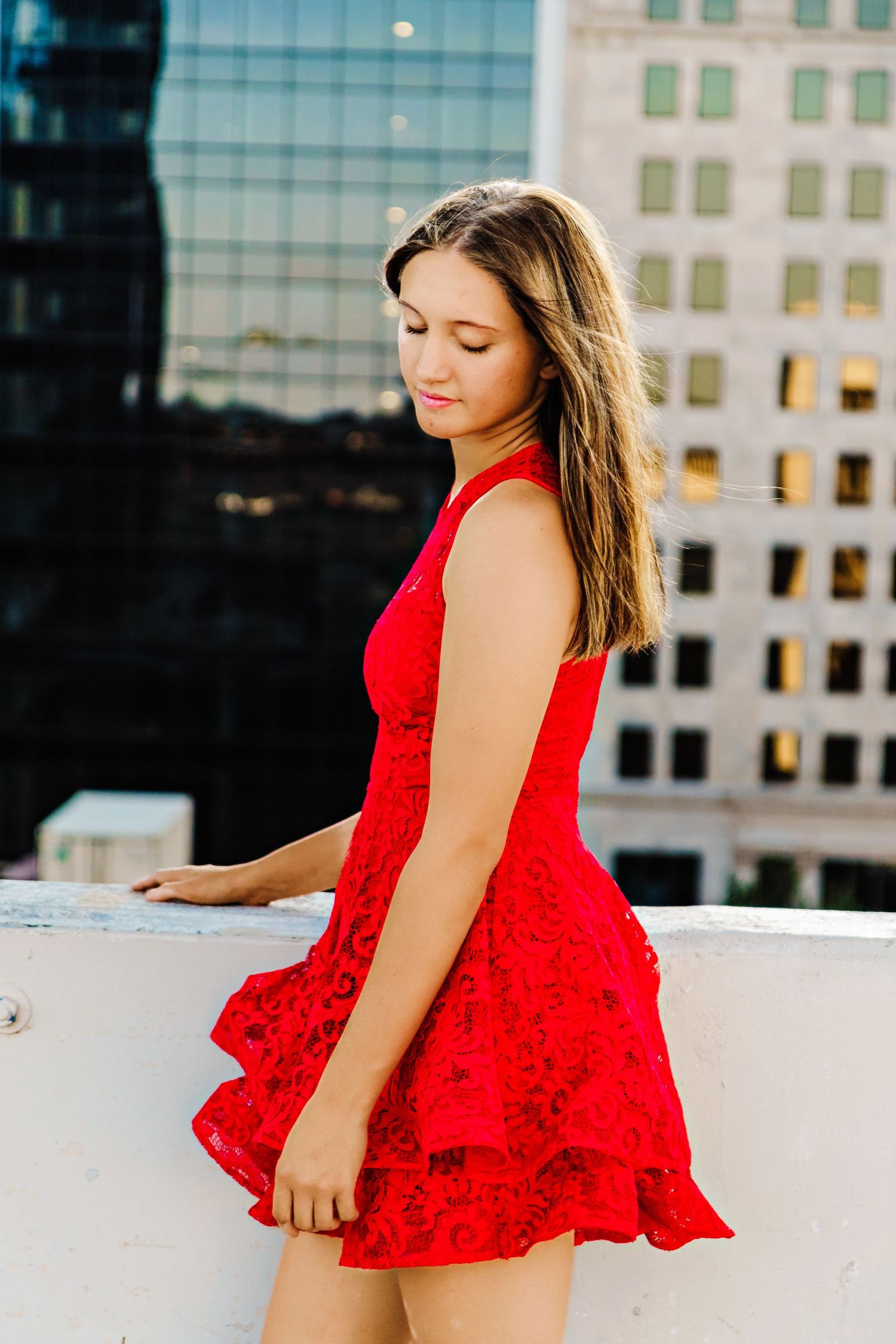 Young woman in a red lace dress poses on a rooftop with city buildings in the background.