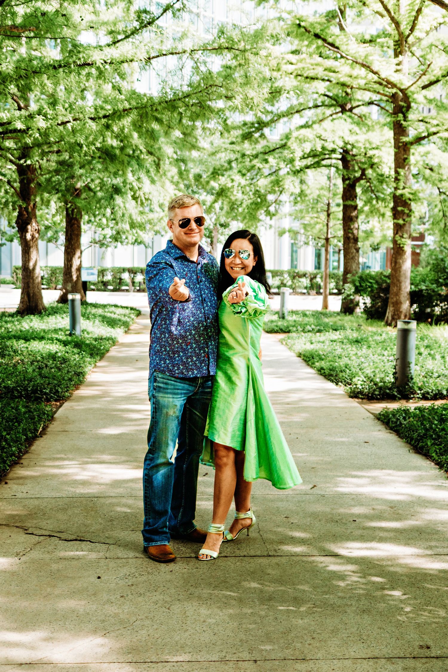 A couple in coordinated blue and green outfits poses playfully in a tree-lined park walkway on a sunny day.
