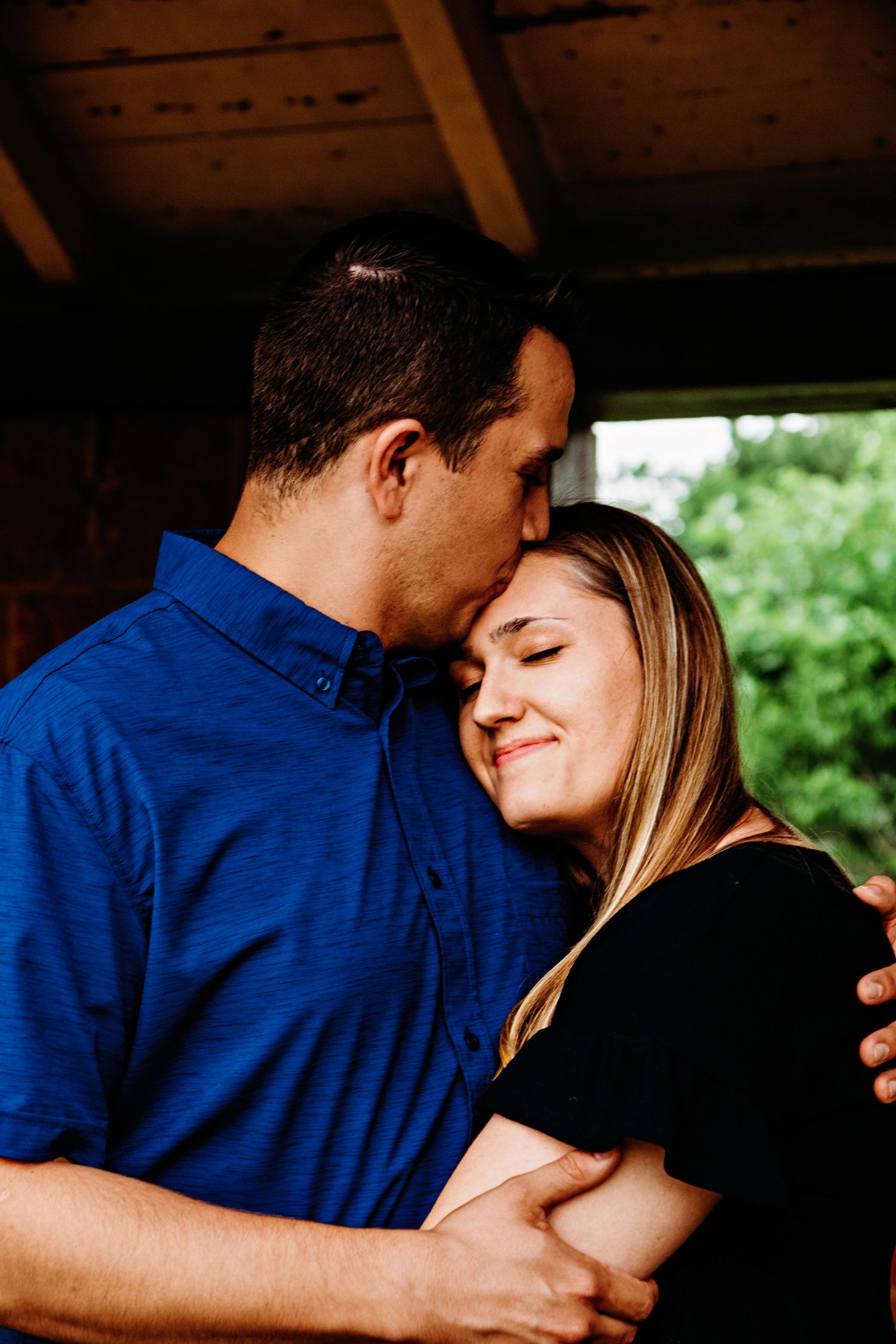 A romantic couple embraces tenderly in a rustic wooden setting with natural lighting and greenery in the background.