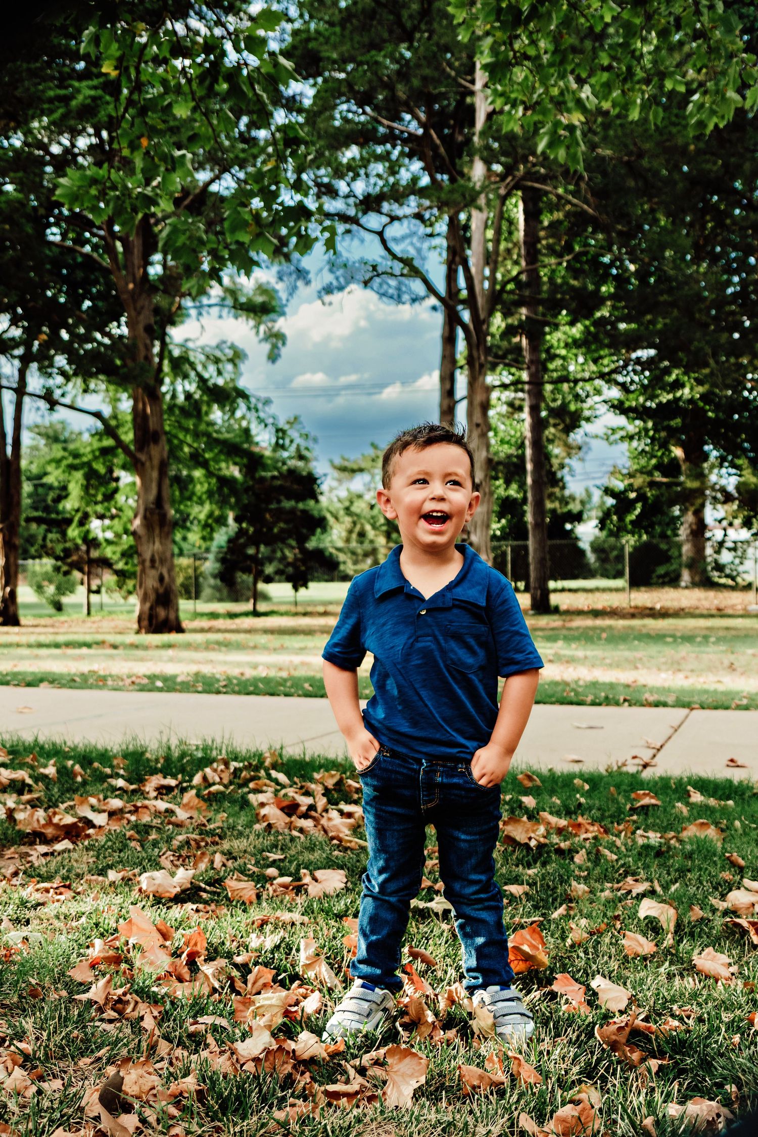 A joyful young boy in a blue polo and jeans stands among fallen autumn leaves in a lush green park.