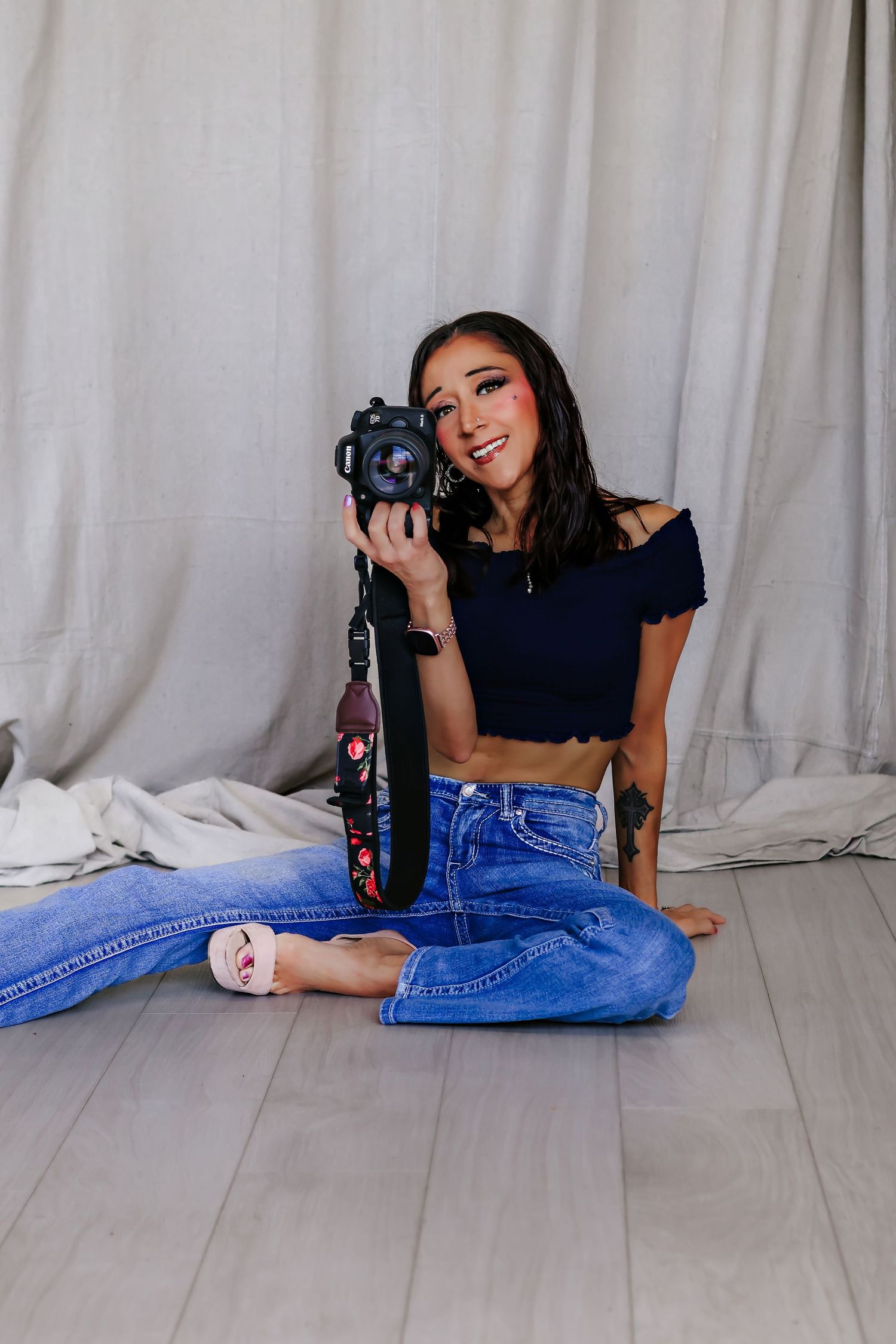 Young woman sitting cross-legged on floor holding DSLR camera, wearing blue crop top and jeans, smiling in studio setting.