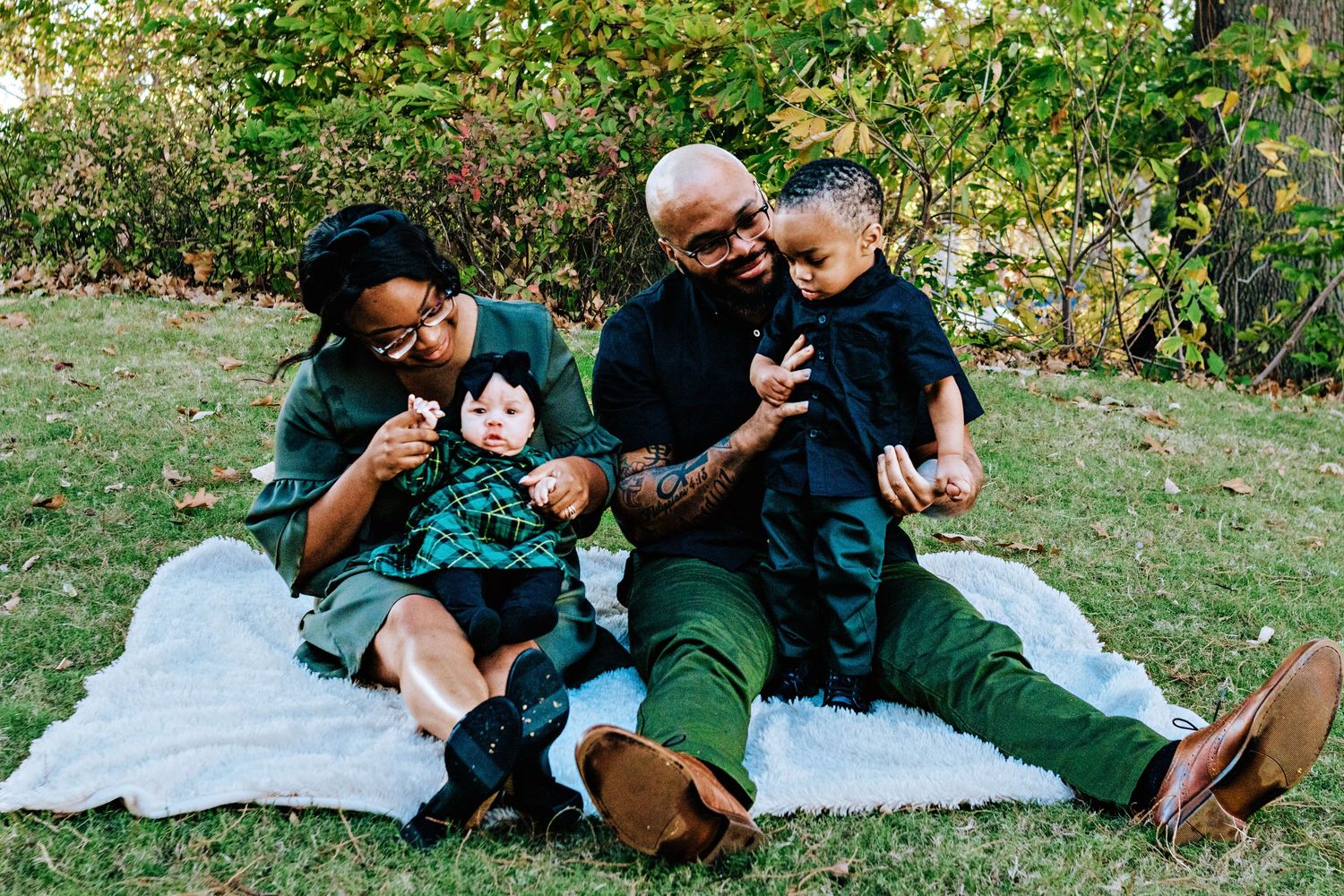 A couple relaxes on a picnic blanket in a grassy area surrounded by trees on a sunny day.