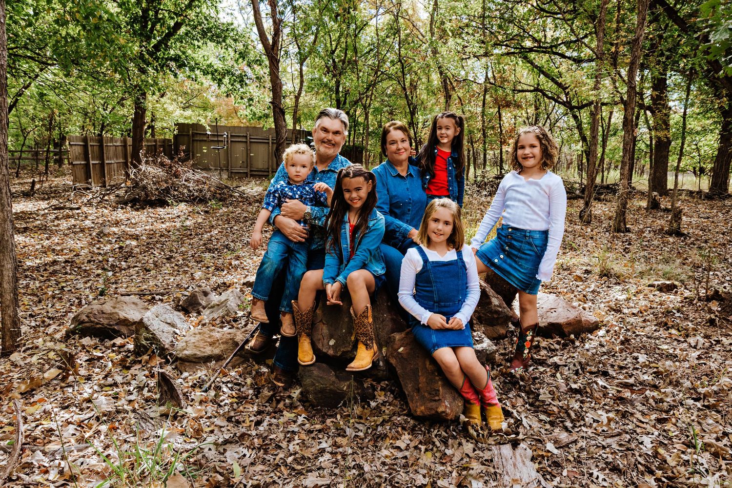 Family of seven posing together on a log in a wooded area, wearing coordinated blue and yellow outfits.