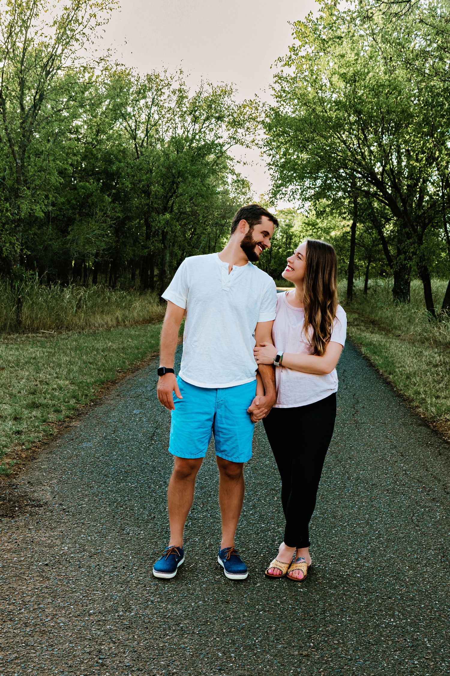 A couple holds hands and smiles at each other on a tree-lined path, man in white tee and blue shorts, woman in black pants.