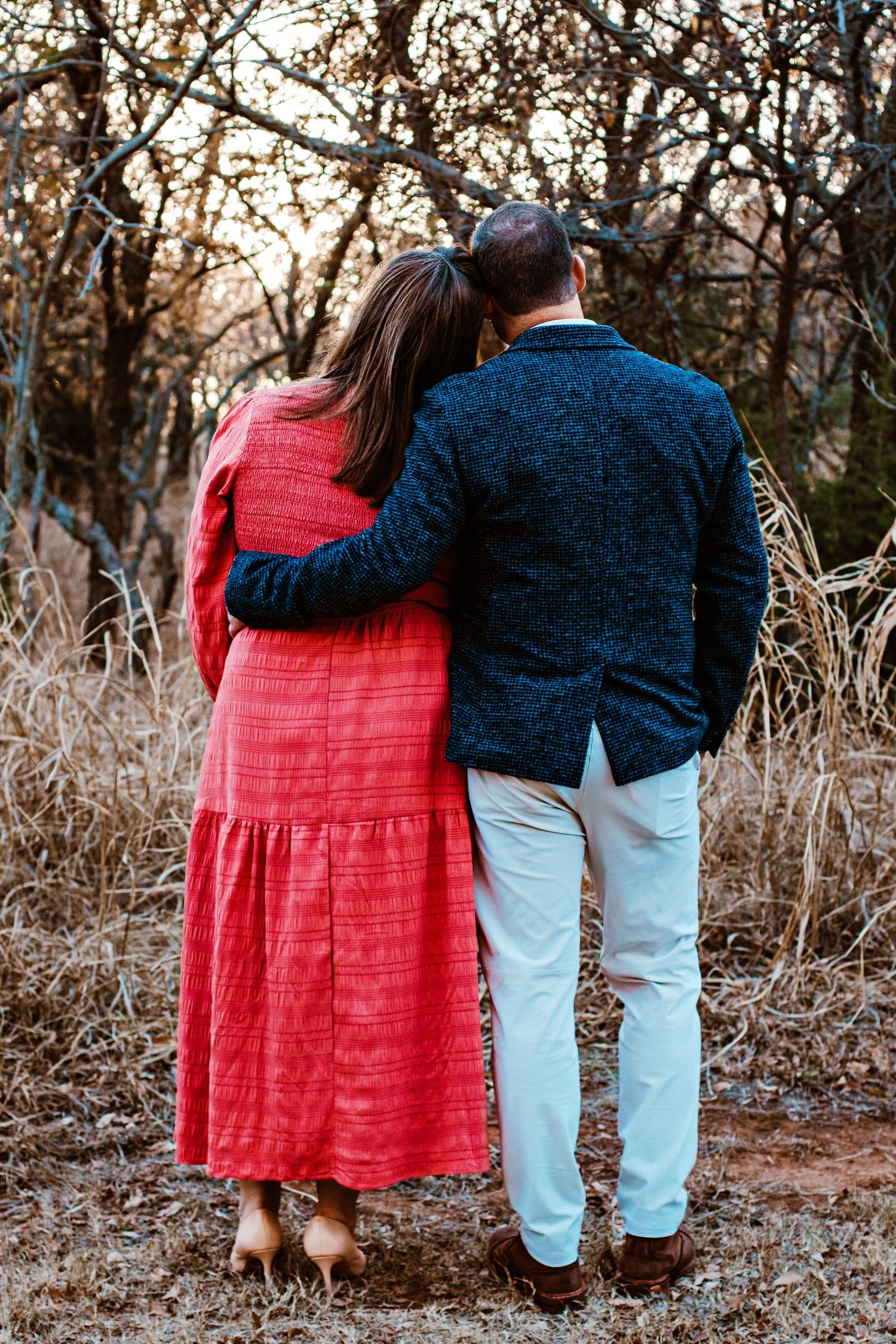A couple in red dress and blue sweater sit together on a rustic bench in an autumn woodland setting.