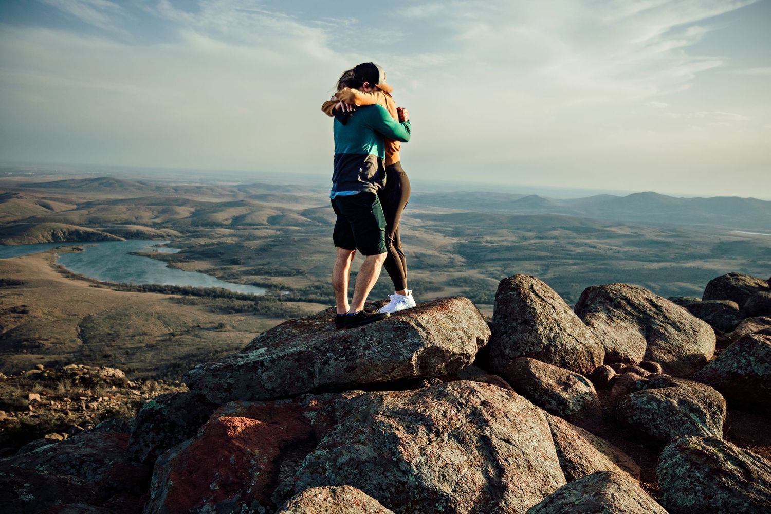 A couple shares a romantic embrace while standing on a mountain summit overlooking a scenic valley landscape at sunset.