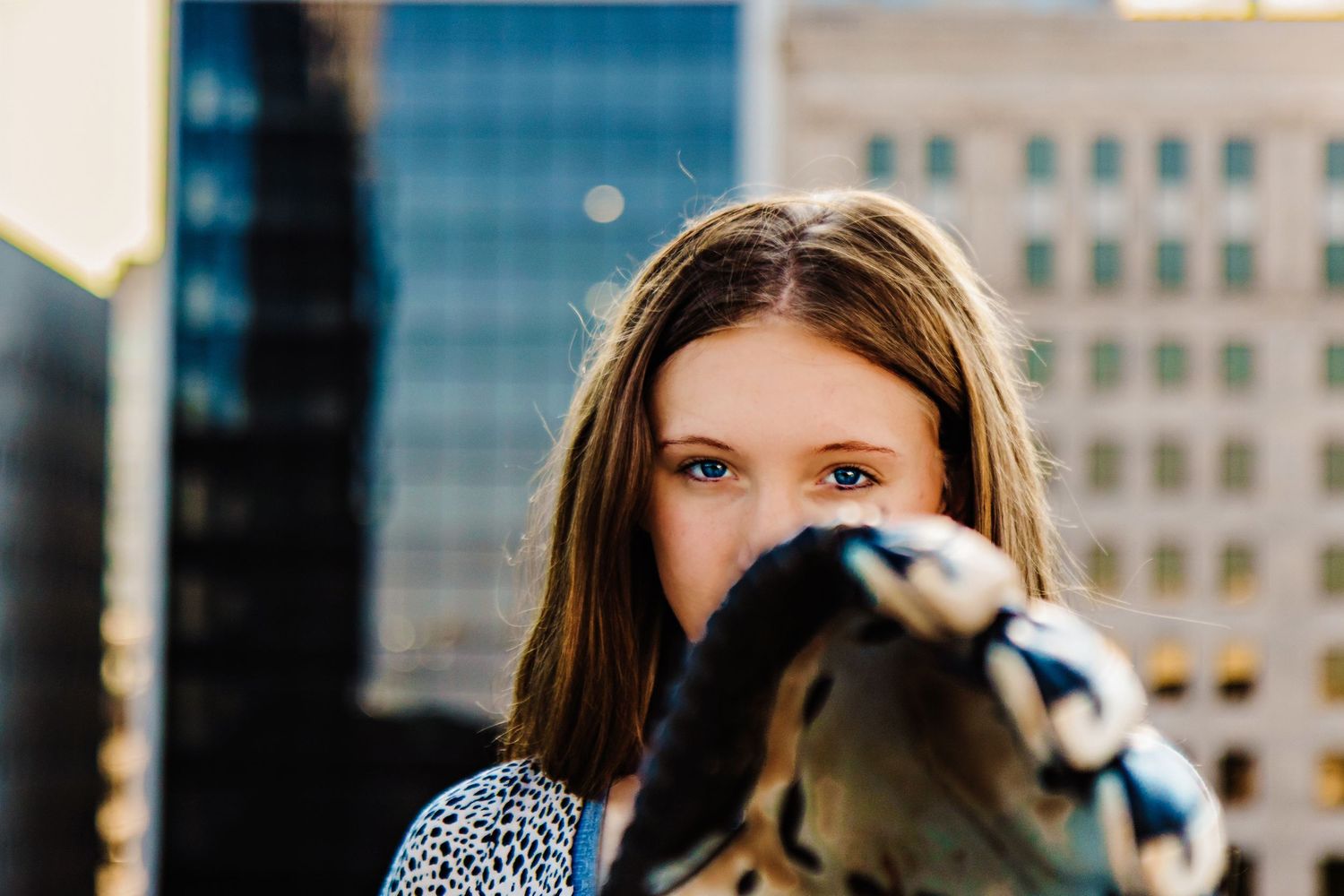 Young woman with brown hair partially covering her face with a glove, standing in front of city buildings.