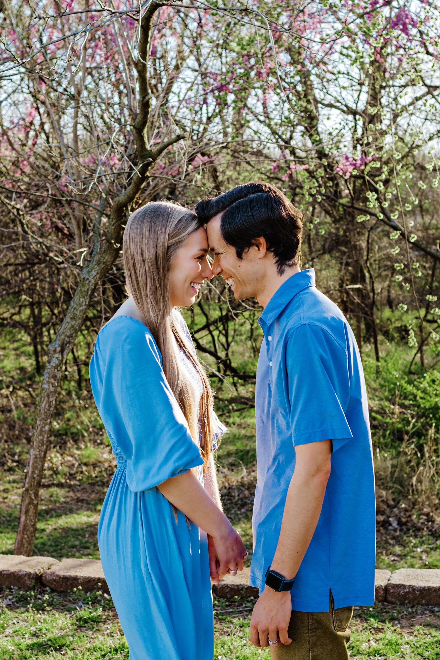A couple in matching blue shirts share a tender moment among blooming cherry blossom trees in a spring garden.