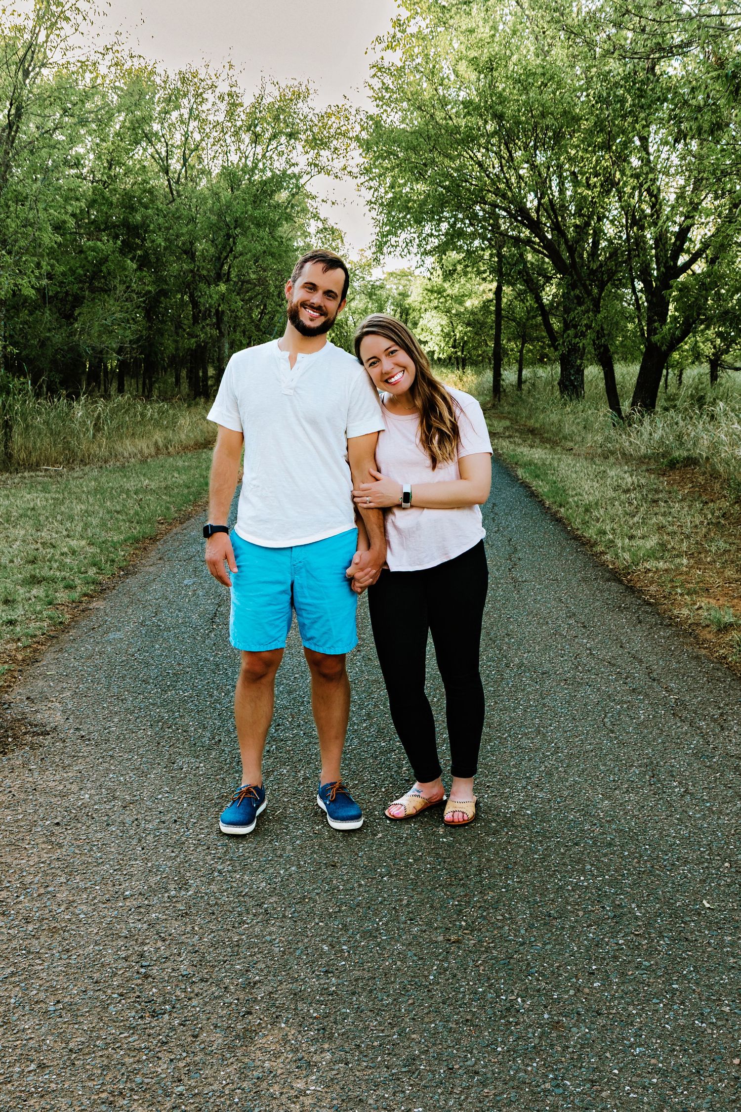 Couple holding hands smiling on a tree-lined road, man in white tee and blue shorts, woman in pink top and black pants.