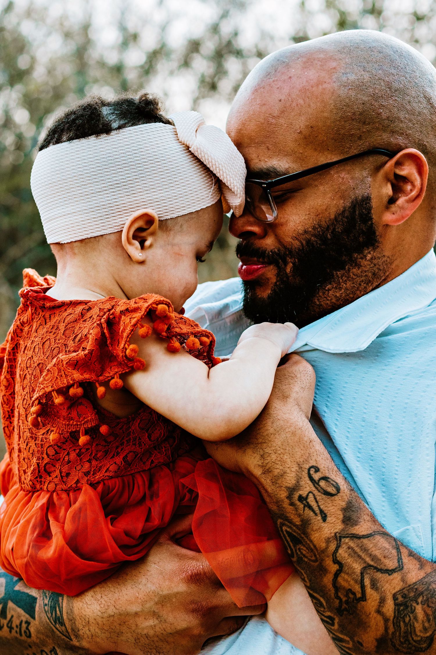 Parent in light blue shirt embraces child wearing orange dress in outdoor setting with natural lighting.