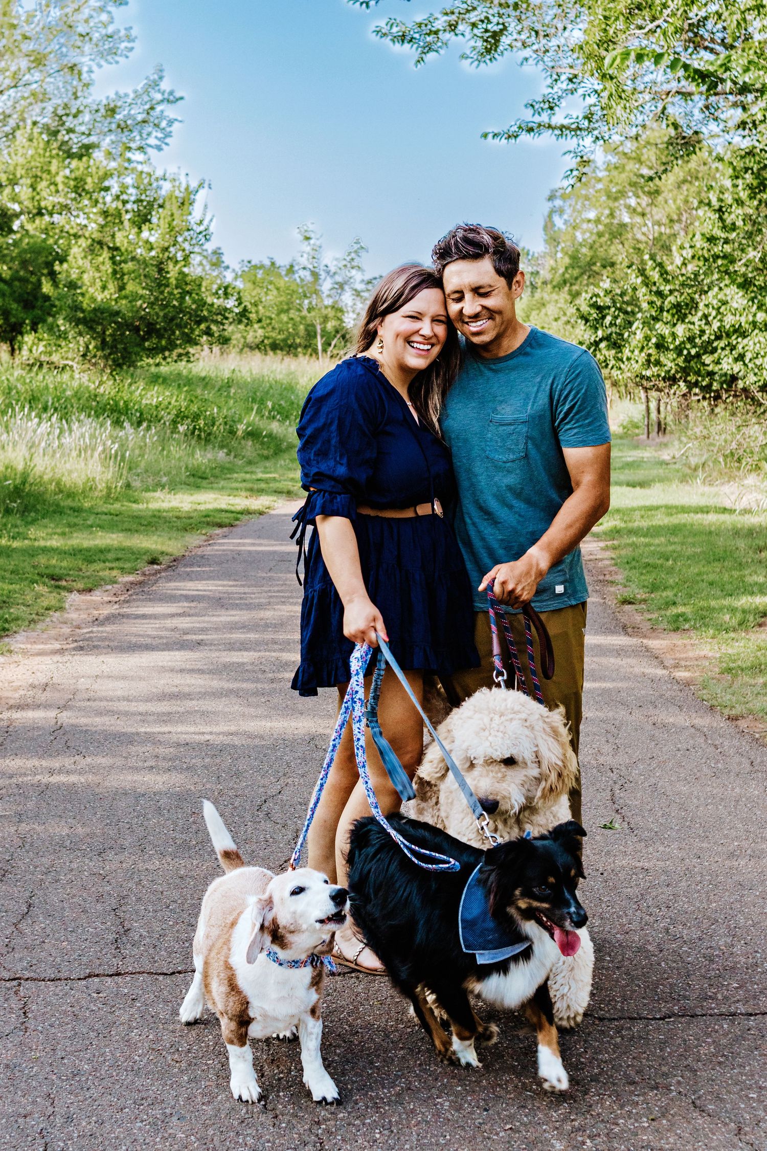 A couple poses with their two dogs on a paved walking trail surrounded by green grass and trees.