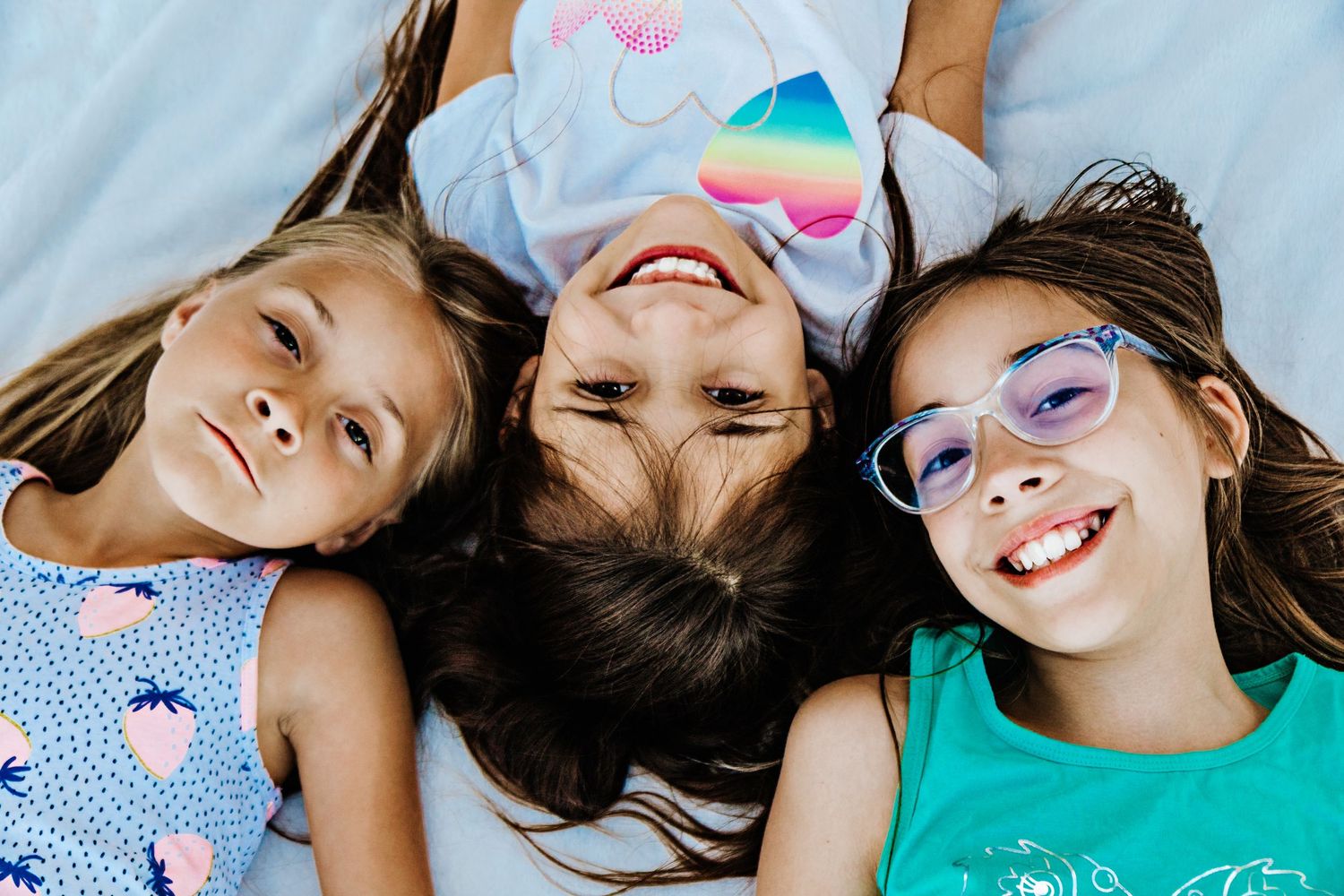 Three smiling girls lying on white bedding, one wearing blue glasses, enjoying a fun playdate together.