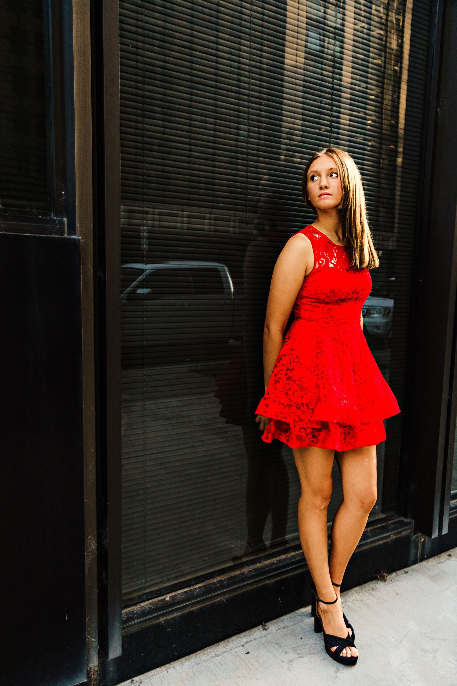 Young blonde woman in a red lace mini dress and black heels posing against a dark glass storefront.