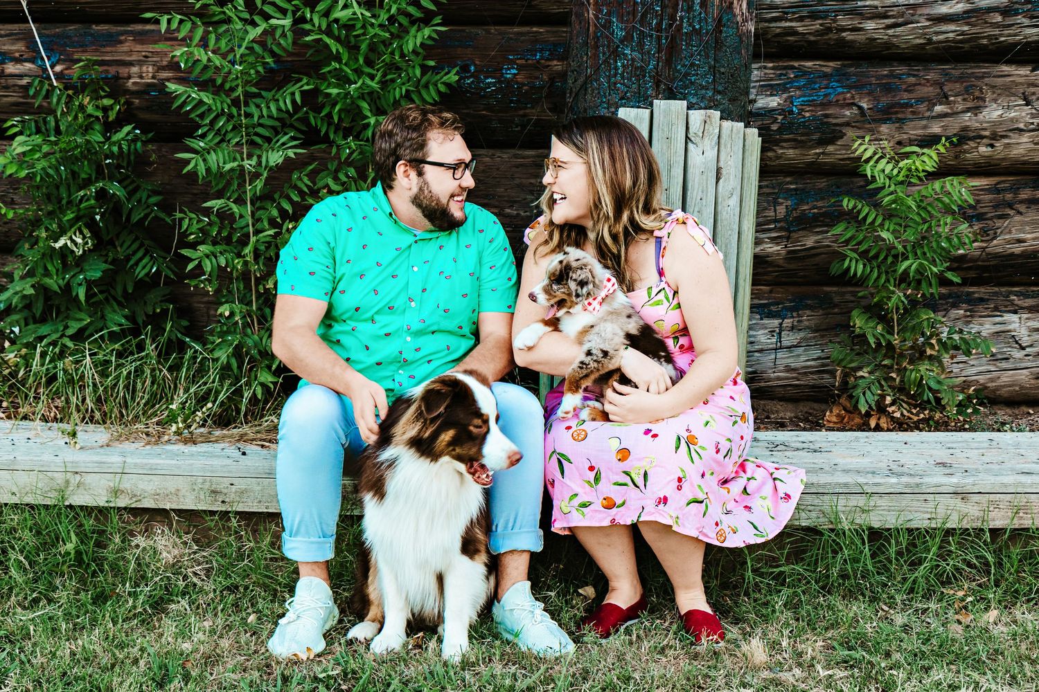 Couple sitting on wooden bench with two dogs, wearing colorful outfits, smiling at each other outdoors.