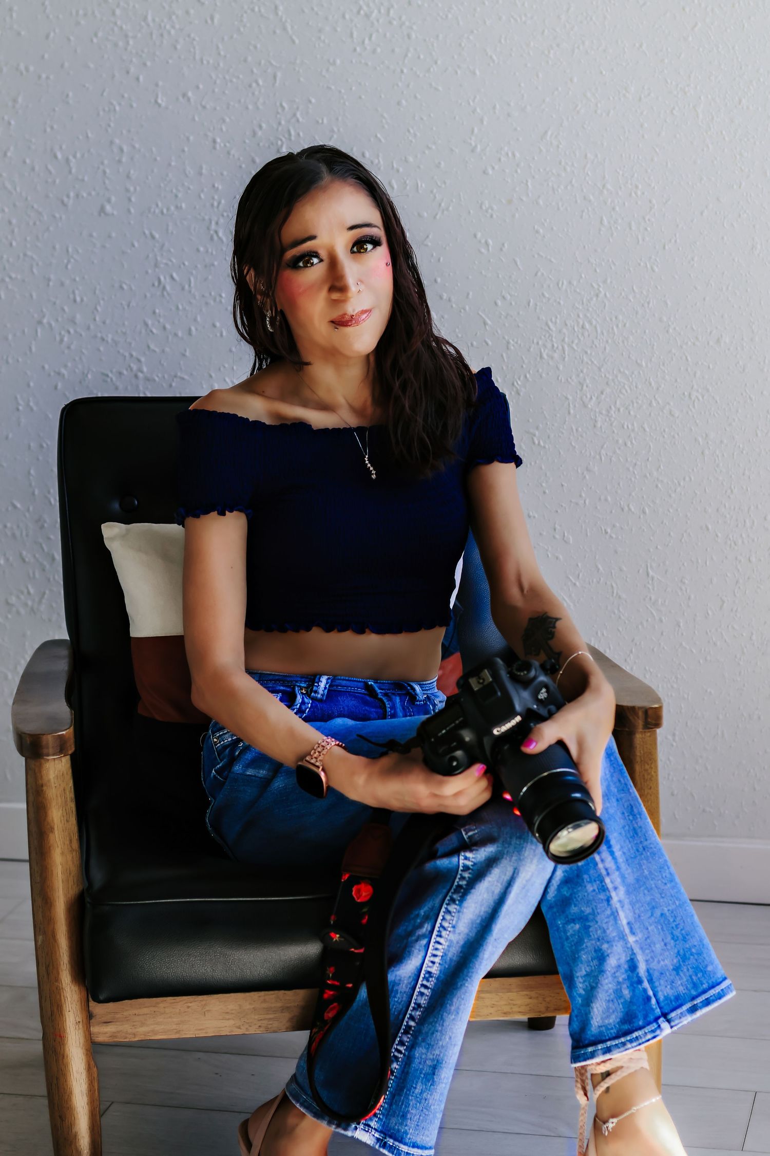 Young woman sitting in a chair holding a DSLR camera, wearing a blue crop top and jeans.