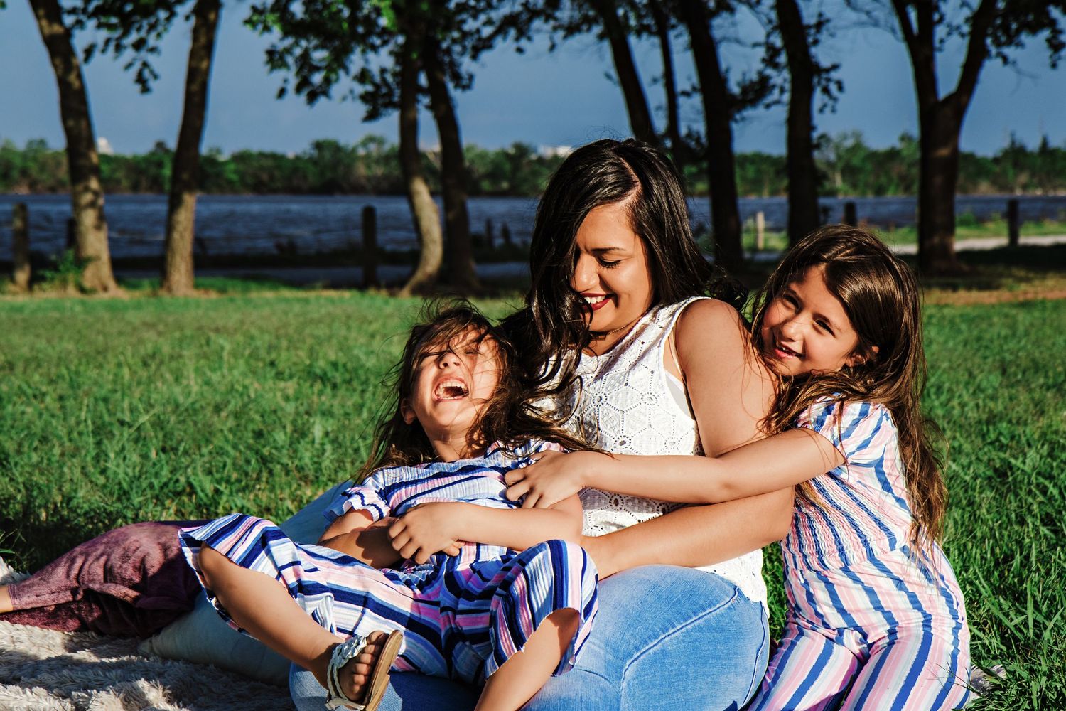 A mother laughs joyfully with her two daughters in striped dresses during a sunny park picnic.