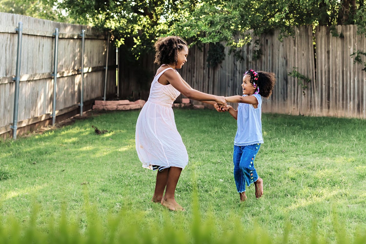 A mother and young daughter joyfully hold hands and spin together in a sunny green backyard.