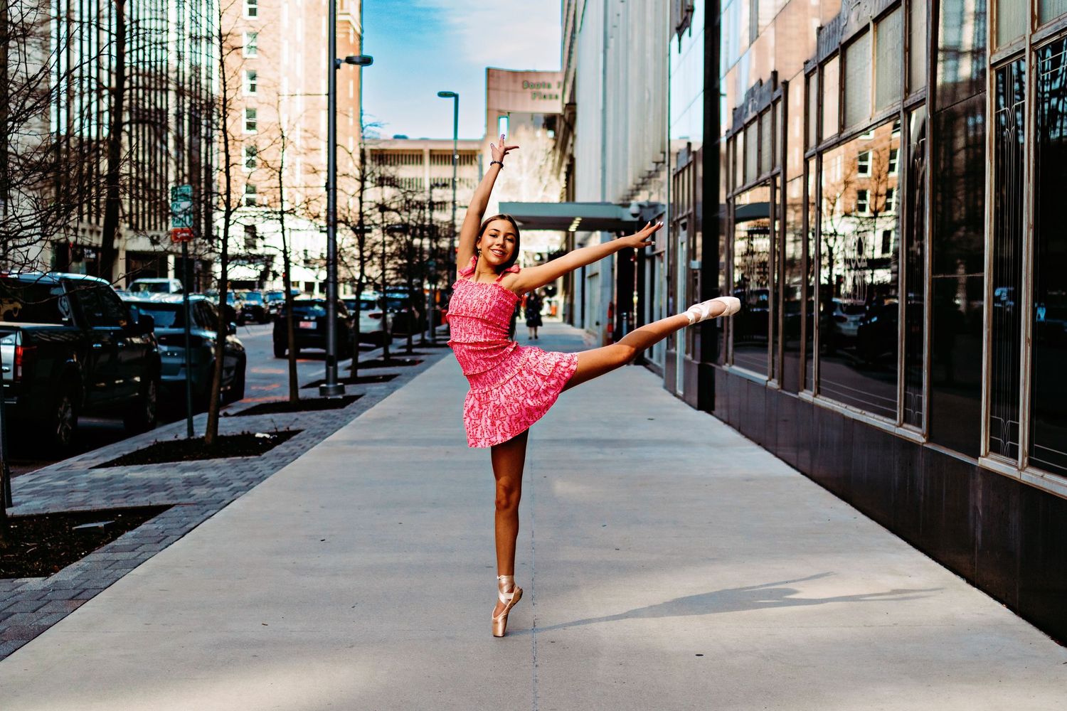 A ballet dancer in a pink dress performs an arabesque pose on a city sidewalk near modern buildings.