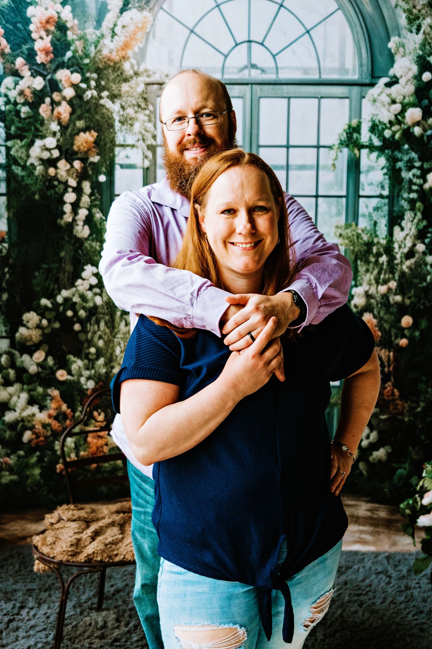 A couple embraces in front of an arched window surrounded by pink and white floral arrangements.
