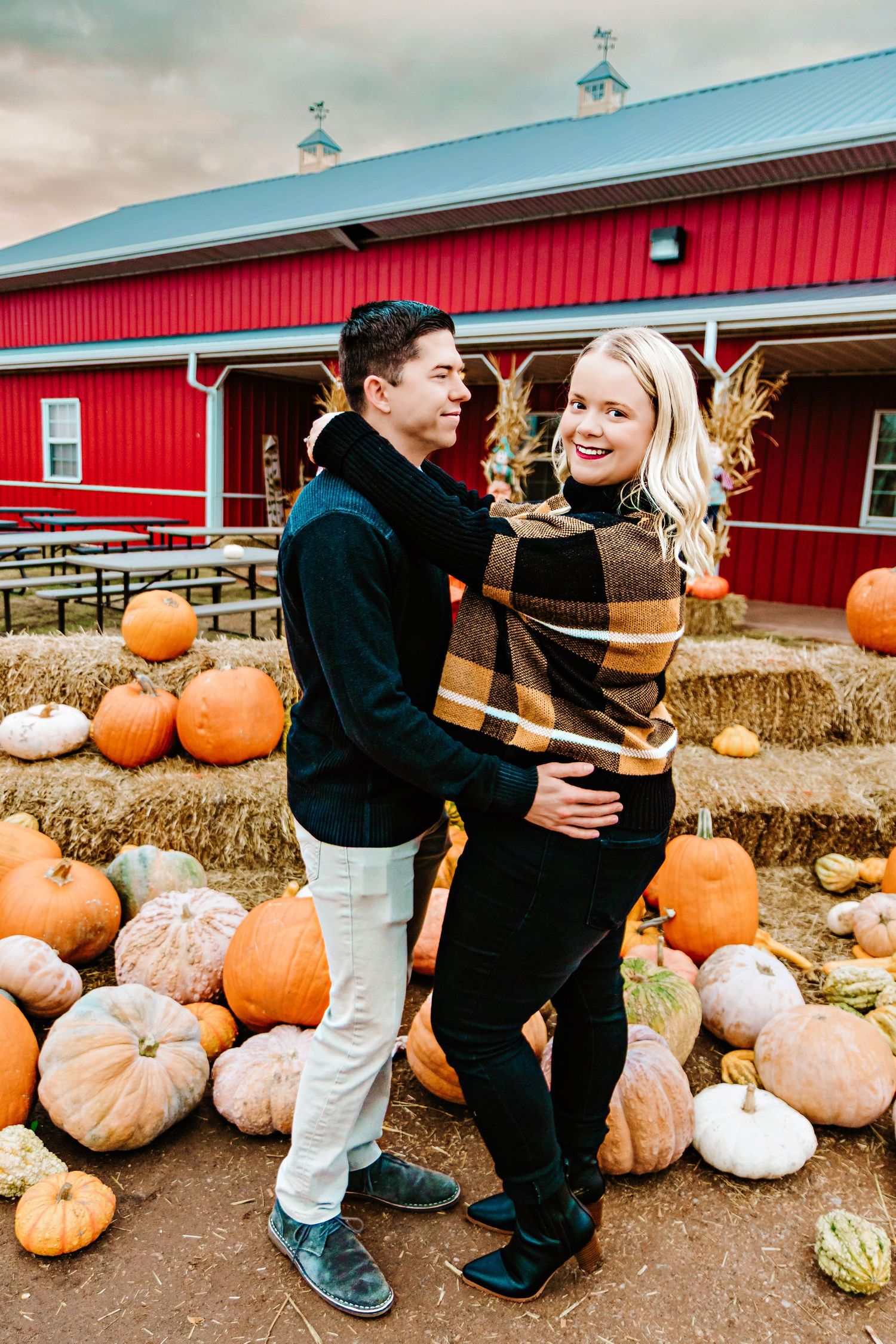 Couple embracing at a fall pumpkin patch with hay bales and a red barn backdrop on a cloudy autumn day.