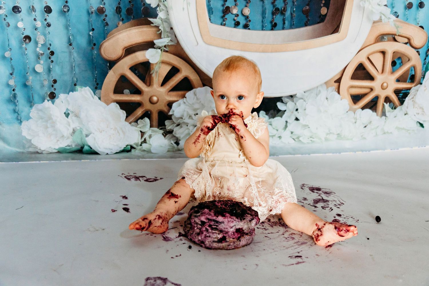 A baby in vintage dress sits on floor with cake during messy cake smash photo session.
