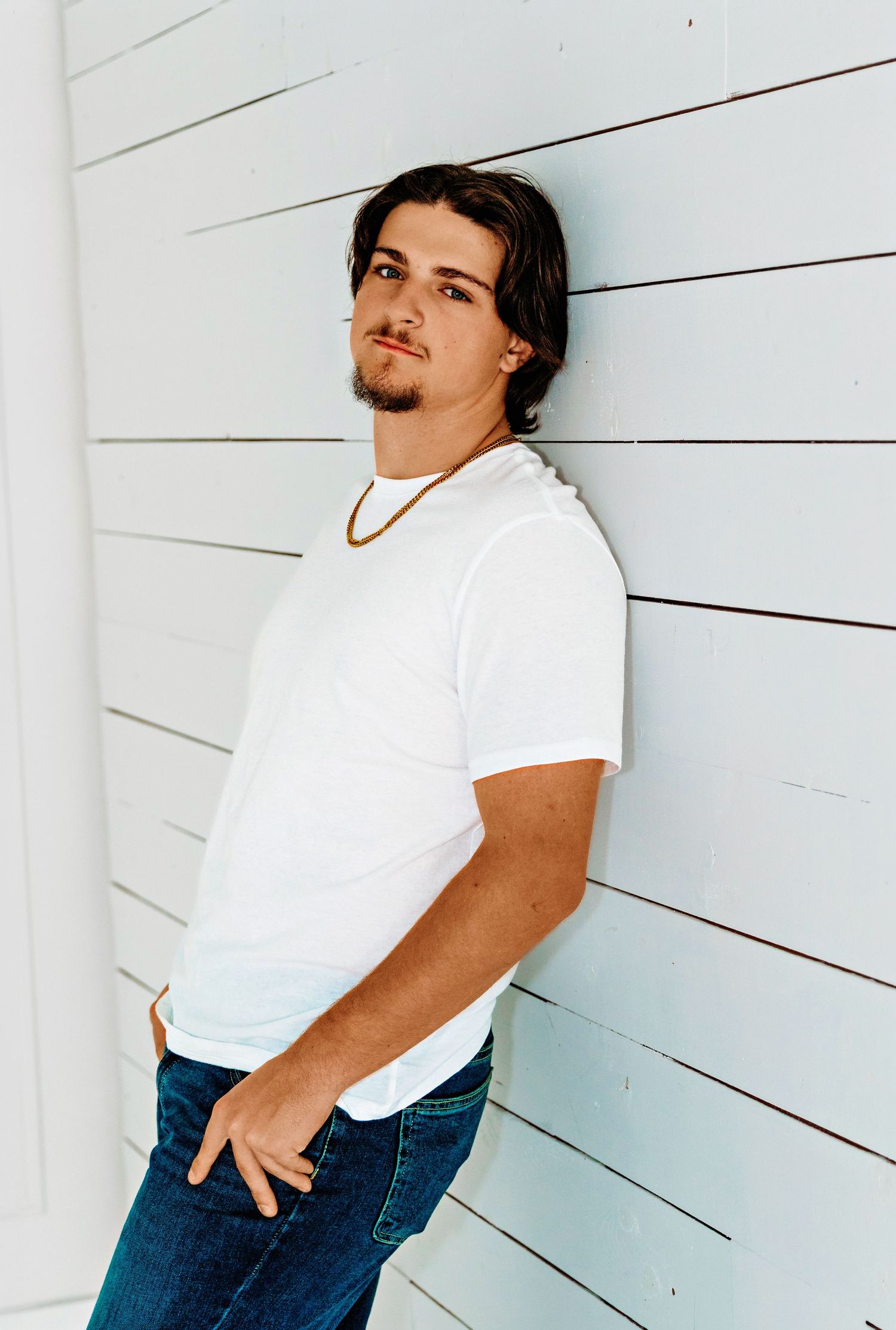 A person in a white t-shirt and jeans poses casually against a white wooden wall in a portrait-style photo.