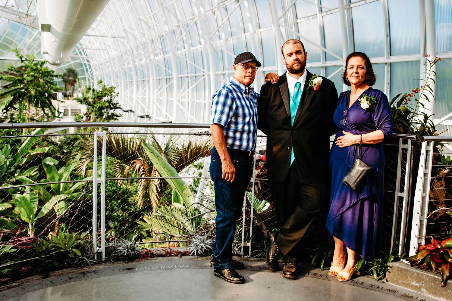 Three people stand together posing for a photo inside a bright greenhouse conservatory with tropical plants.