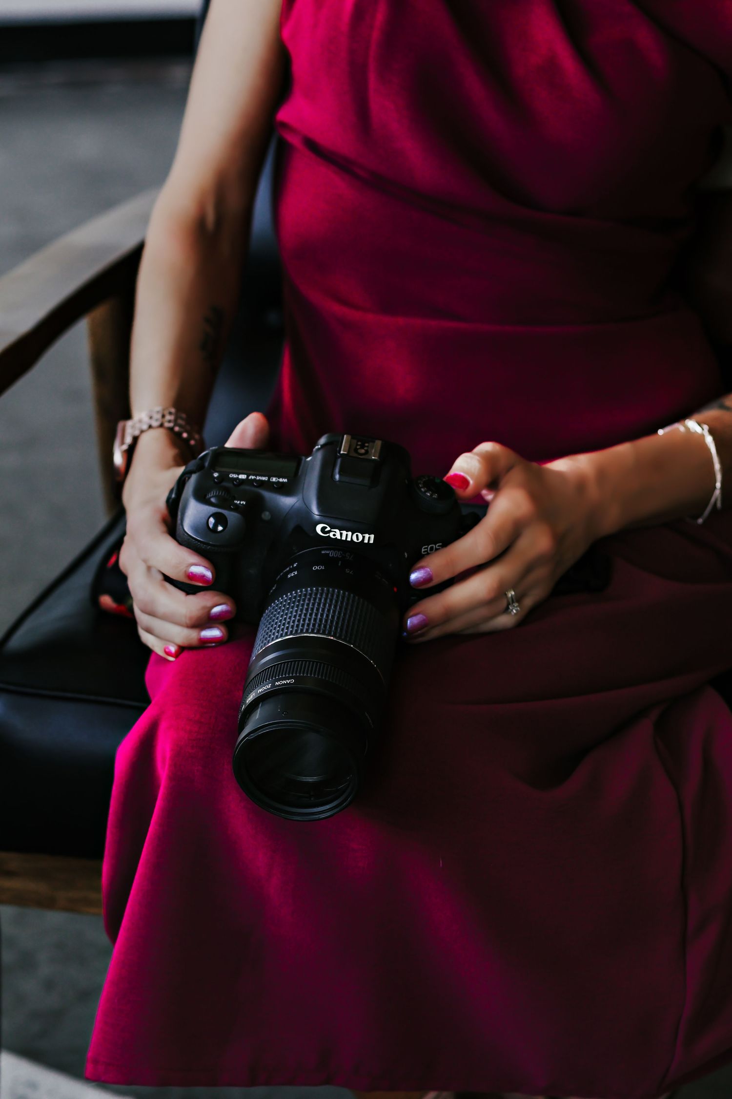 Woman in red dress holding a Canon DSLR camera with telephoto lens, wearing red nail polish and jewelry.