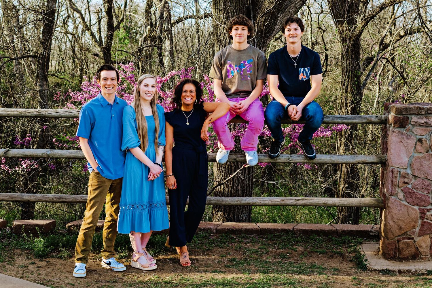 A group of people sit together on a rustic wooden fence in an outdoor wooded setting during daytime.