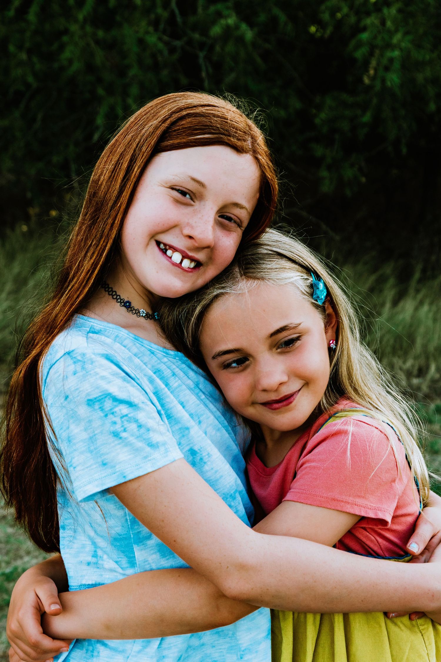 Two young girls hugging and smiling outdoors, one with red hair in a blue tie-dye shirt, the other blonde in a pink shirt.